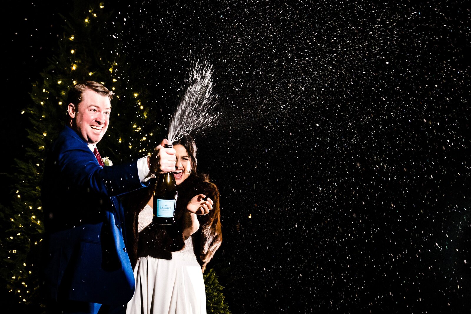 bride and groom pop champagne and the spray is backlit on the dark sky behind them