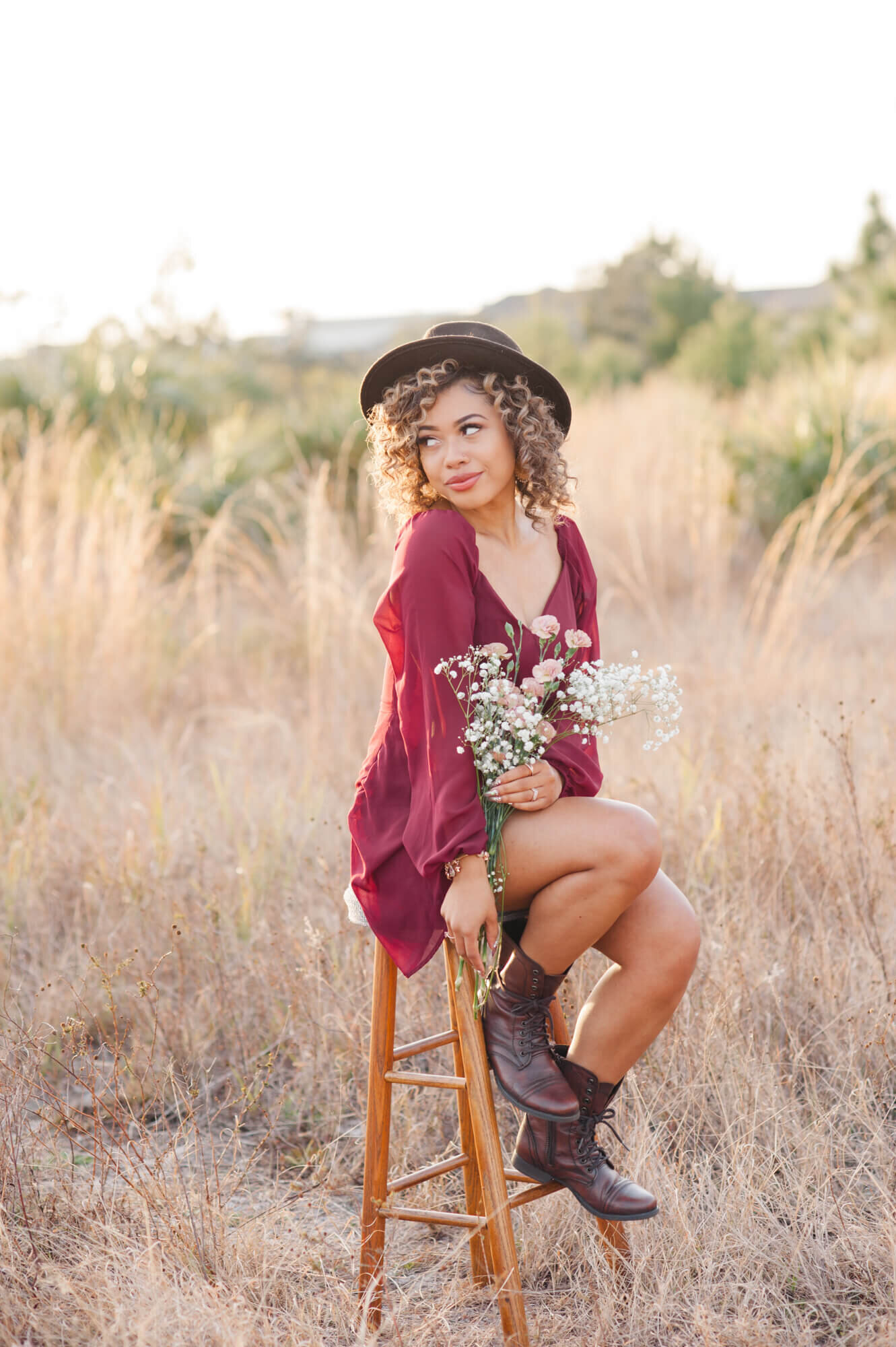 Stunning senior girl with curly hair holding a floral bouquet and sitting on a barstool in a tall grass field at sunset