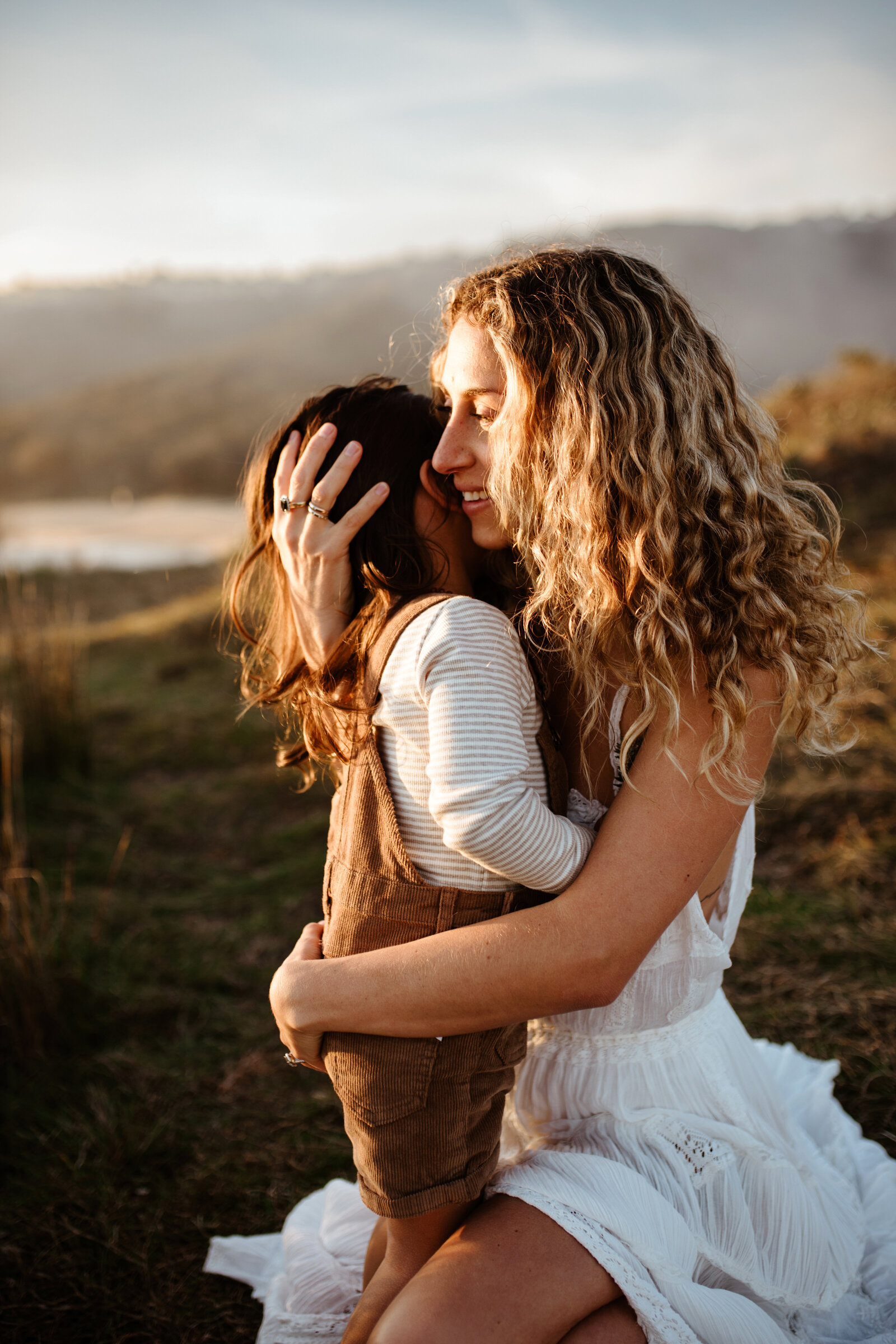 A woman with curly hair in a white dress hugs a child in brown overalls and a long-sleeve shirt while sitting on grass in an outdoor setting.