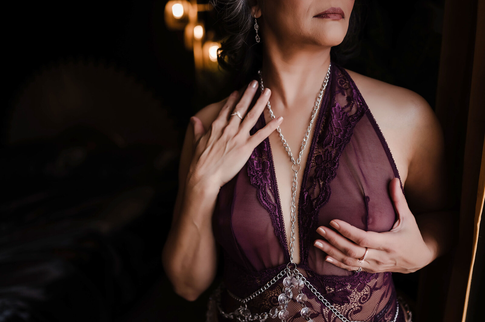 A woman in purple lingerie holds her neck and chest while standing in a window
