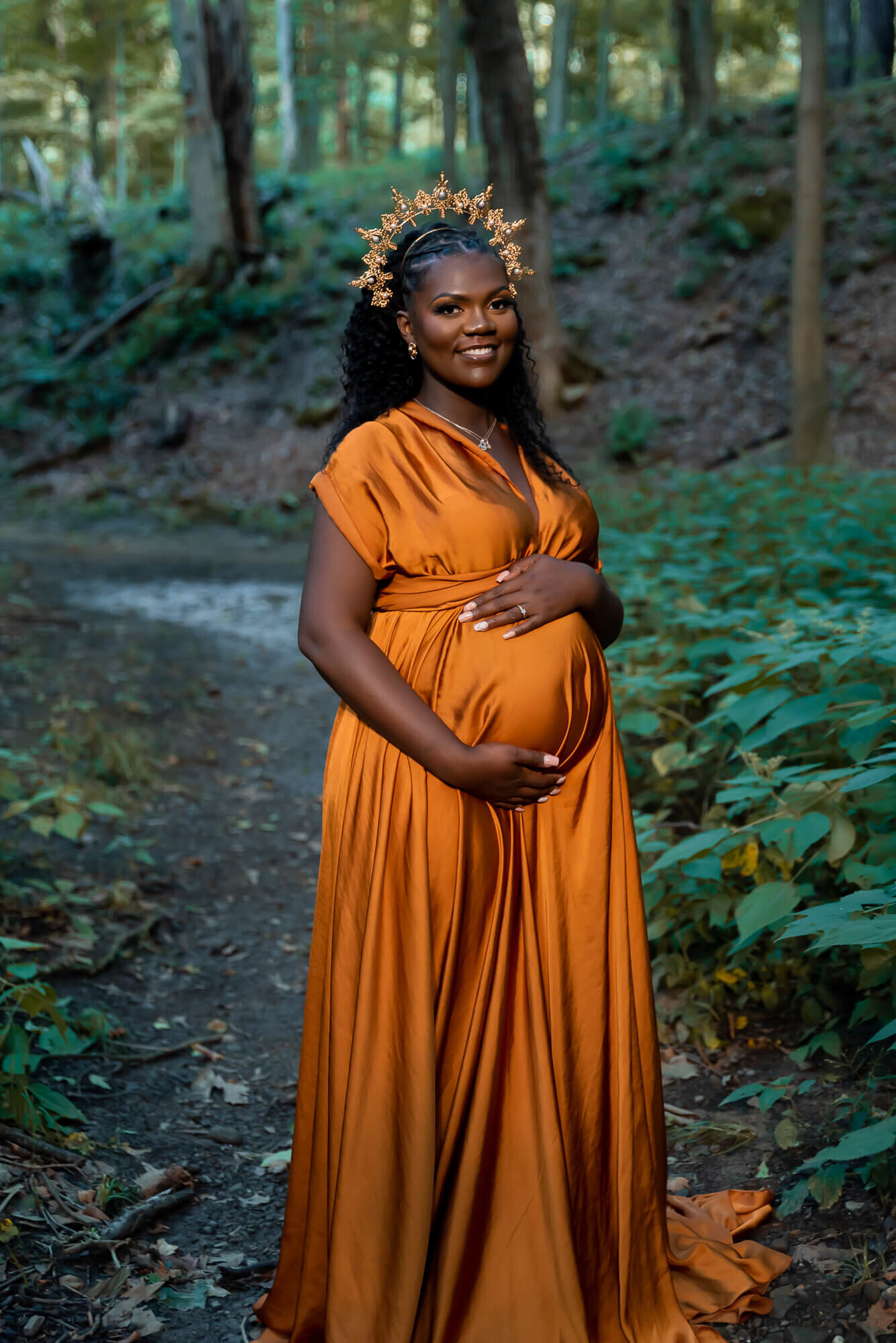A pregnant woman in an orange satin gown and a gold crown stands in a lush forest, holding her belly with a radiant smile.