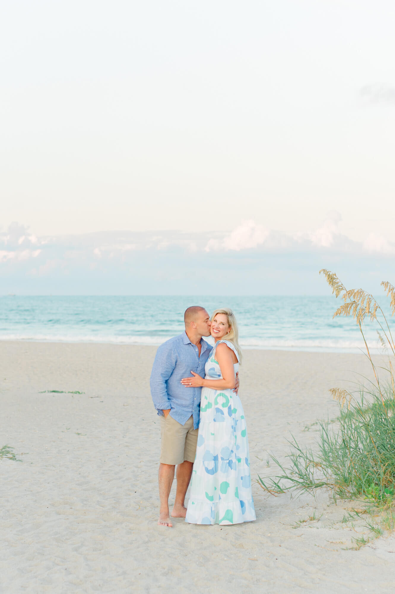 Husband and wife standing on the beach near the dunes and kissing her on the cheek