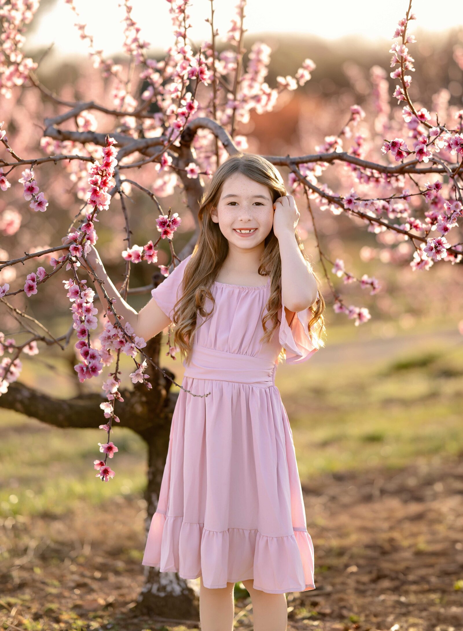 Girl standing by pink peach blossom tree at Ellijay peach orchards near Atlanta, GA. Family photo taken by Jenni Nikki Photography near Atlanta, GA.
