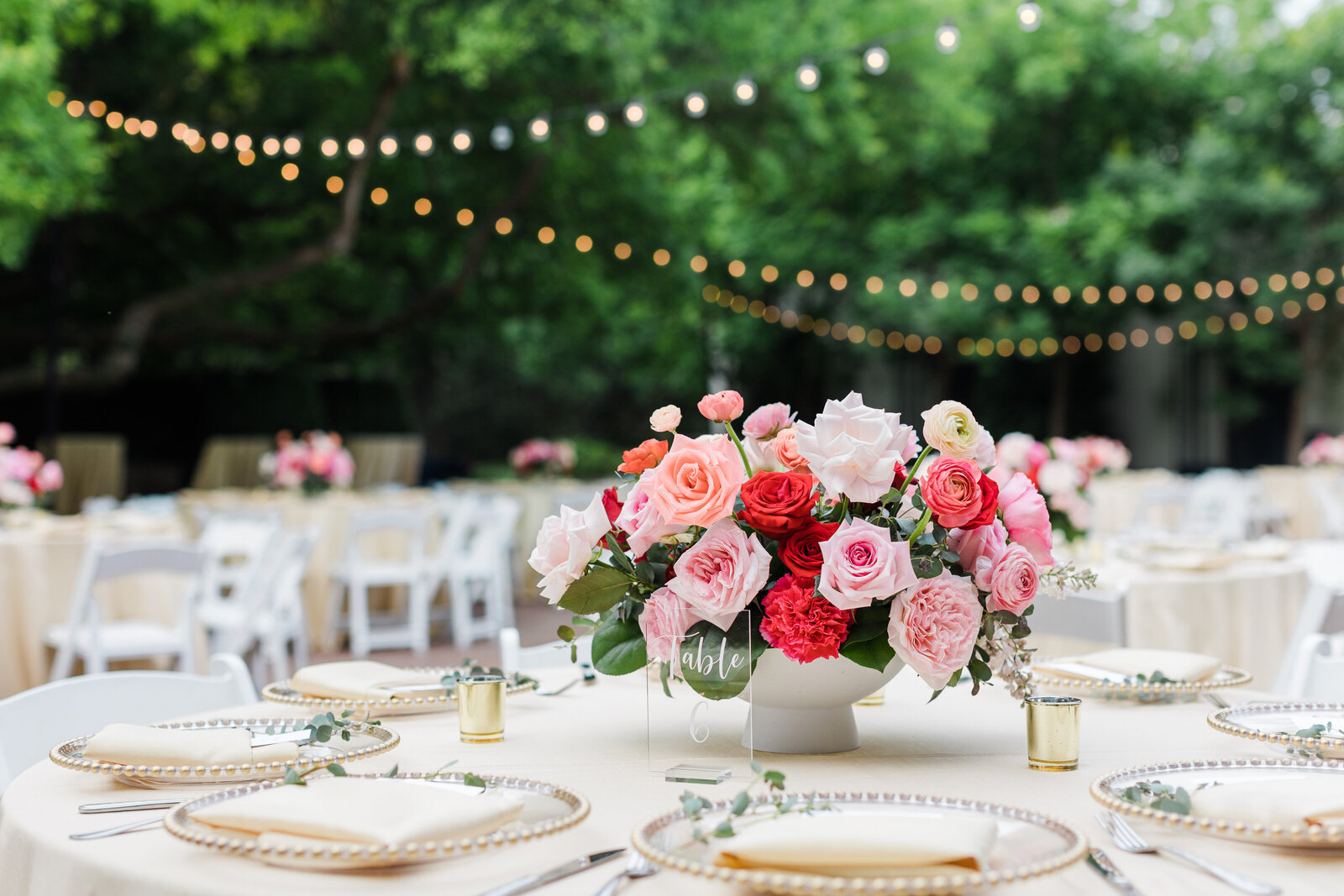 A wedding centerpiece of bold pink and blue flowers sits on a table of plates, napkins, and glassware. It's springtime, so there are vibrant green trees in the background and softly lit cafe lights making tiny little orbs in the background.
