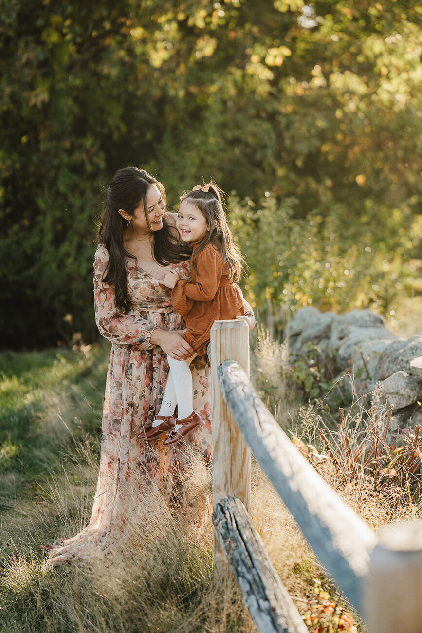 mom smiles with toddler girl sitting on split rail fence