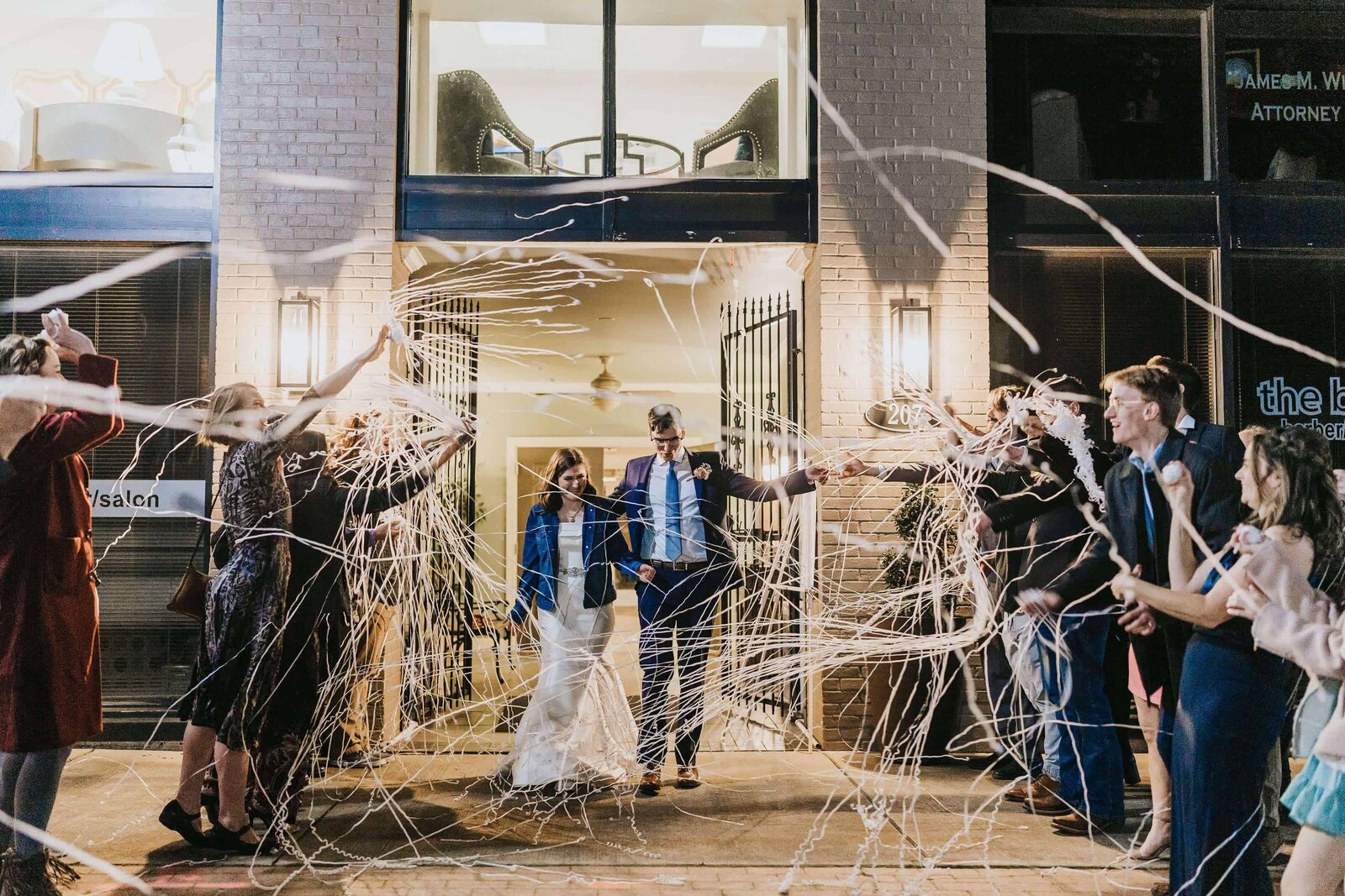 A bride and groom walk out of The Norton Building in Ruston, Louisiana at night, smiling as guests on both sides throw white streamers in celebration. The scene is festive, with people cheering and taking photos.