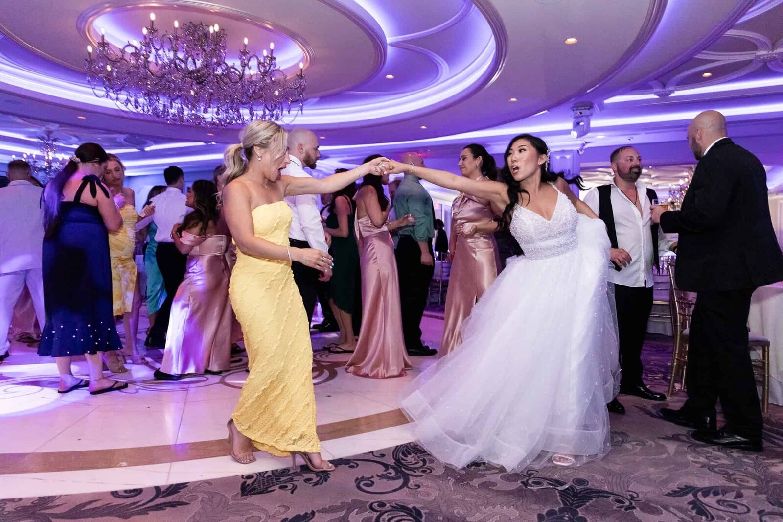 a bride dances with a friend in a yellow dress under an opulent chandelier in a purple lit ballroom
