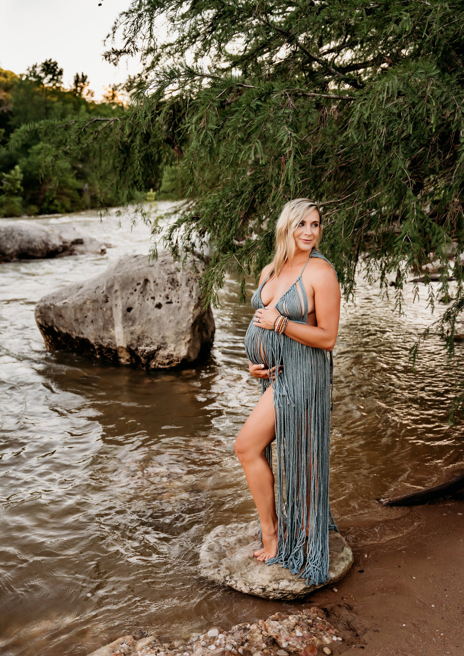 Maternity Photographer, a pregnant woman in a lacy dress stands on stones on the river's bank