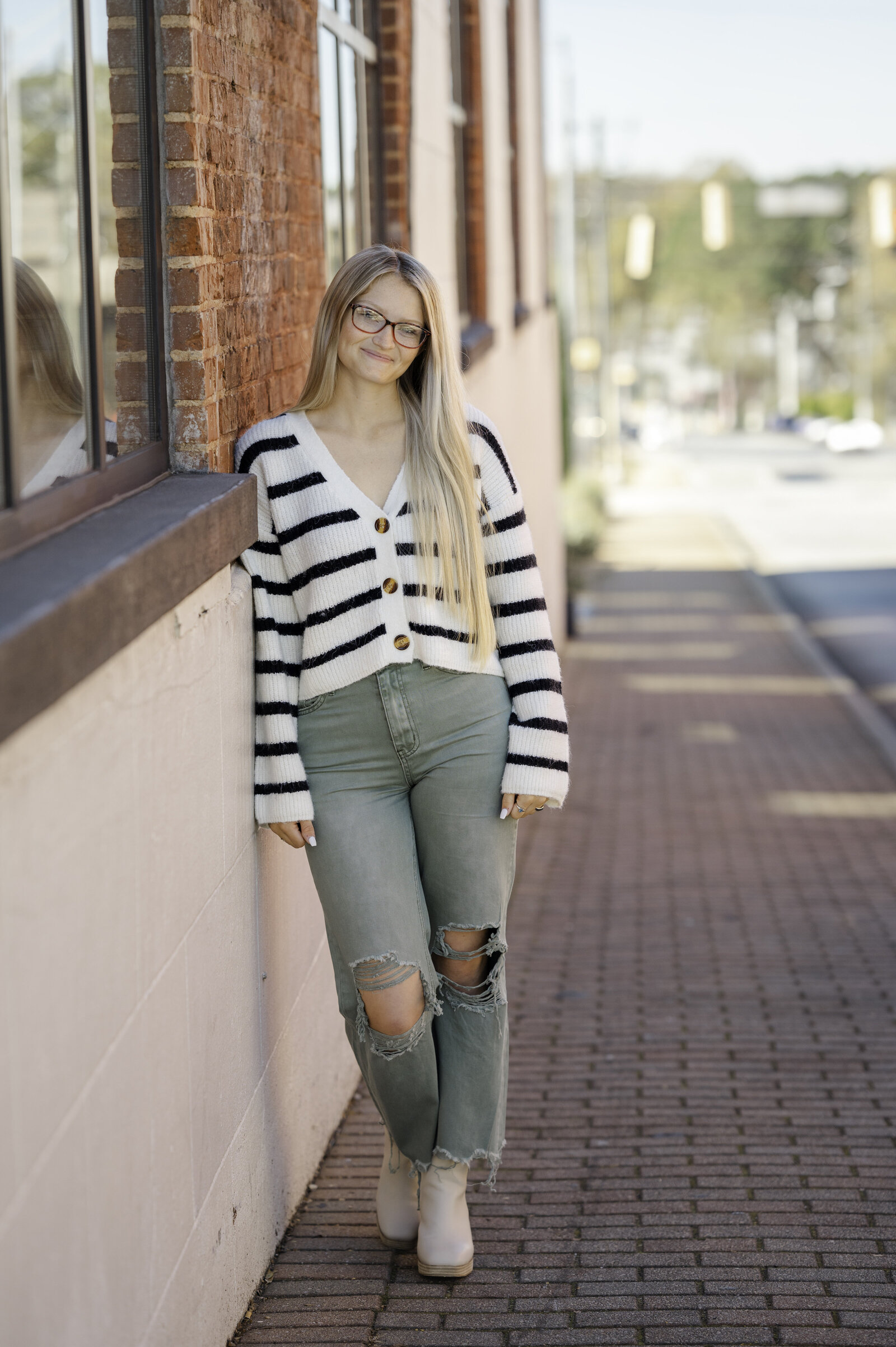 Blonde girl leans against a brick wall wearing army green pants and a black and white stripped sweater