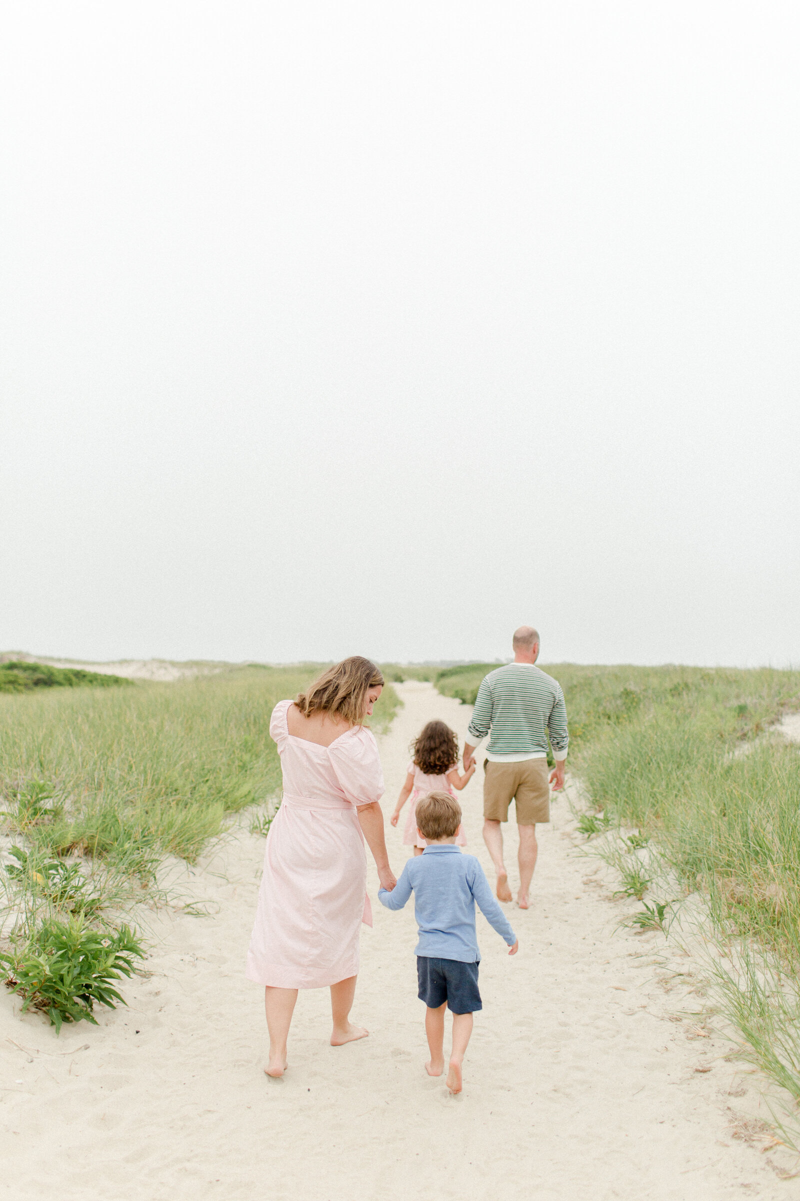 A family of four is walking along a sandy path at the beach and holding hands during photo session with Boston photographer Corinne Isabelle