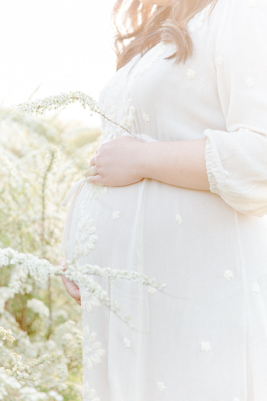 a close up image of a pregnant mother's belly while she is wearing a soft, white maternity gown and standing amongst white wildflowers during photo session with Boston family photographer Corinne Isabelle