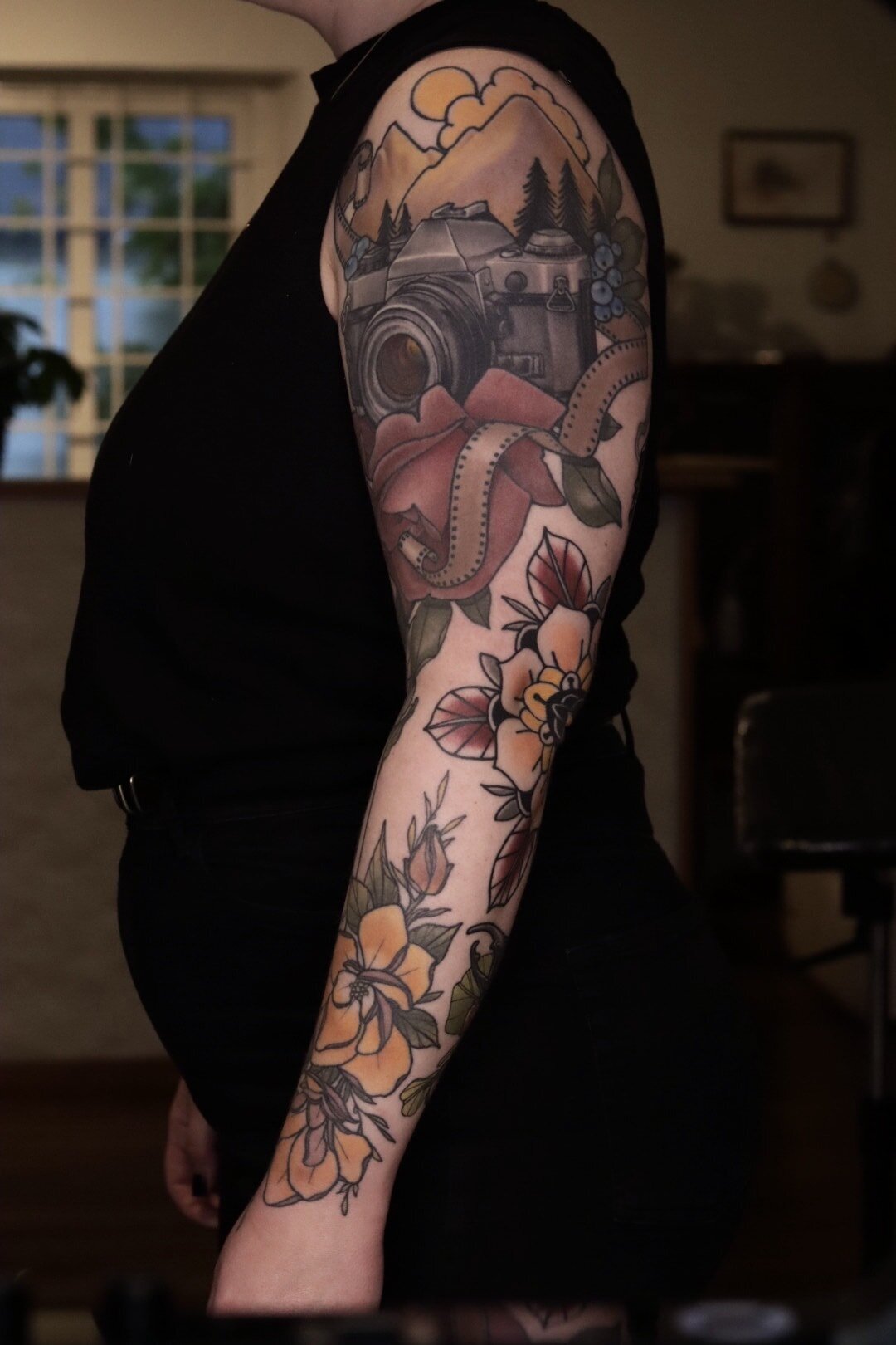 Neo traditional tatuering på helarm med kamera, växter och berg.