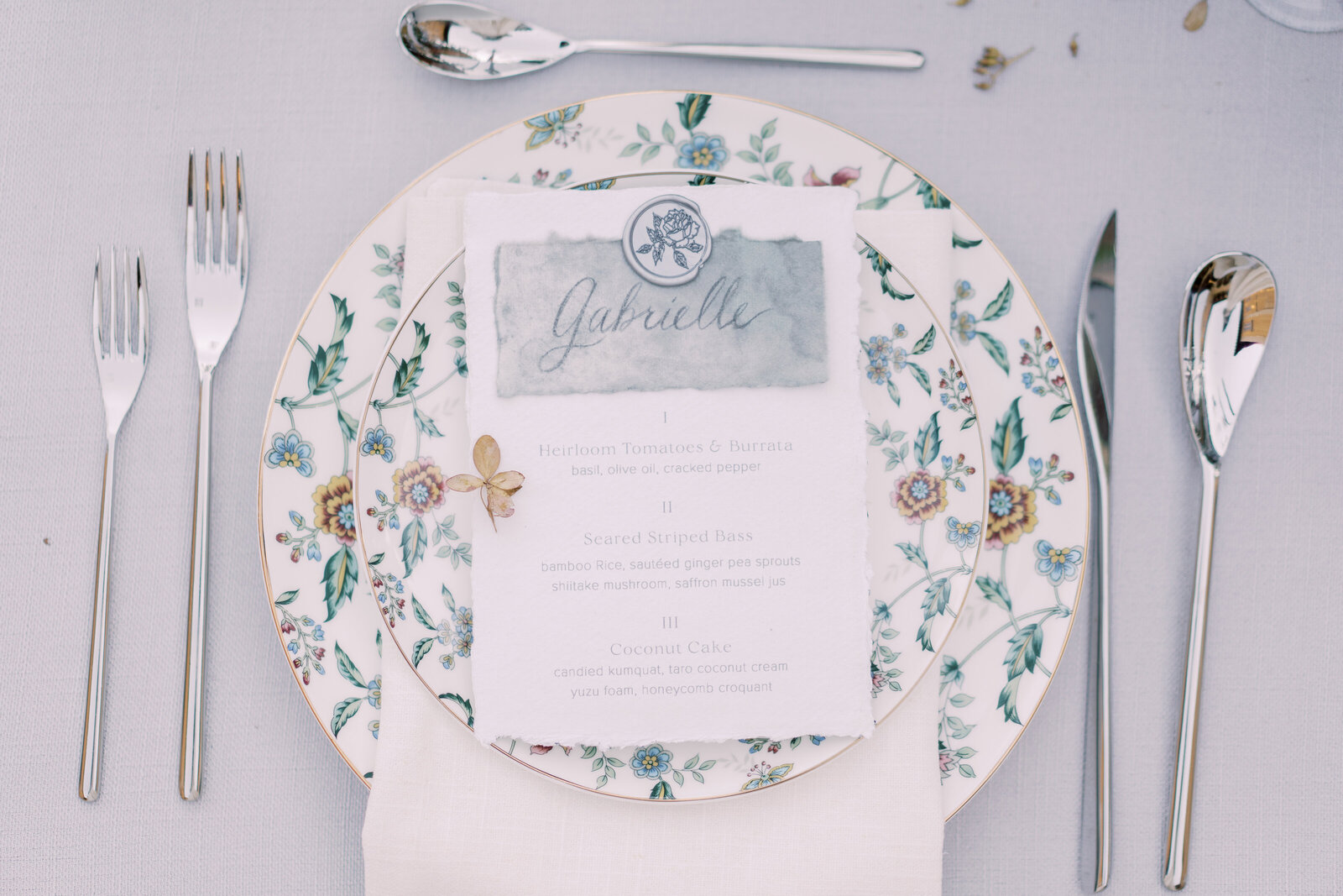 Menu place card on white card stock with gray cursive font atop green floral plates with silver utensils.