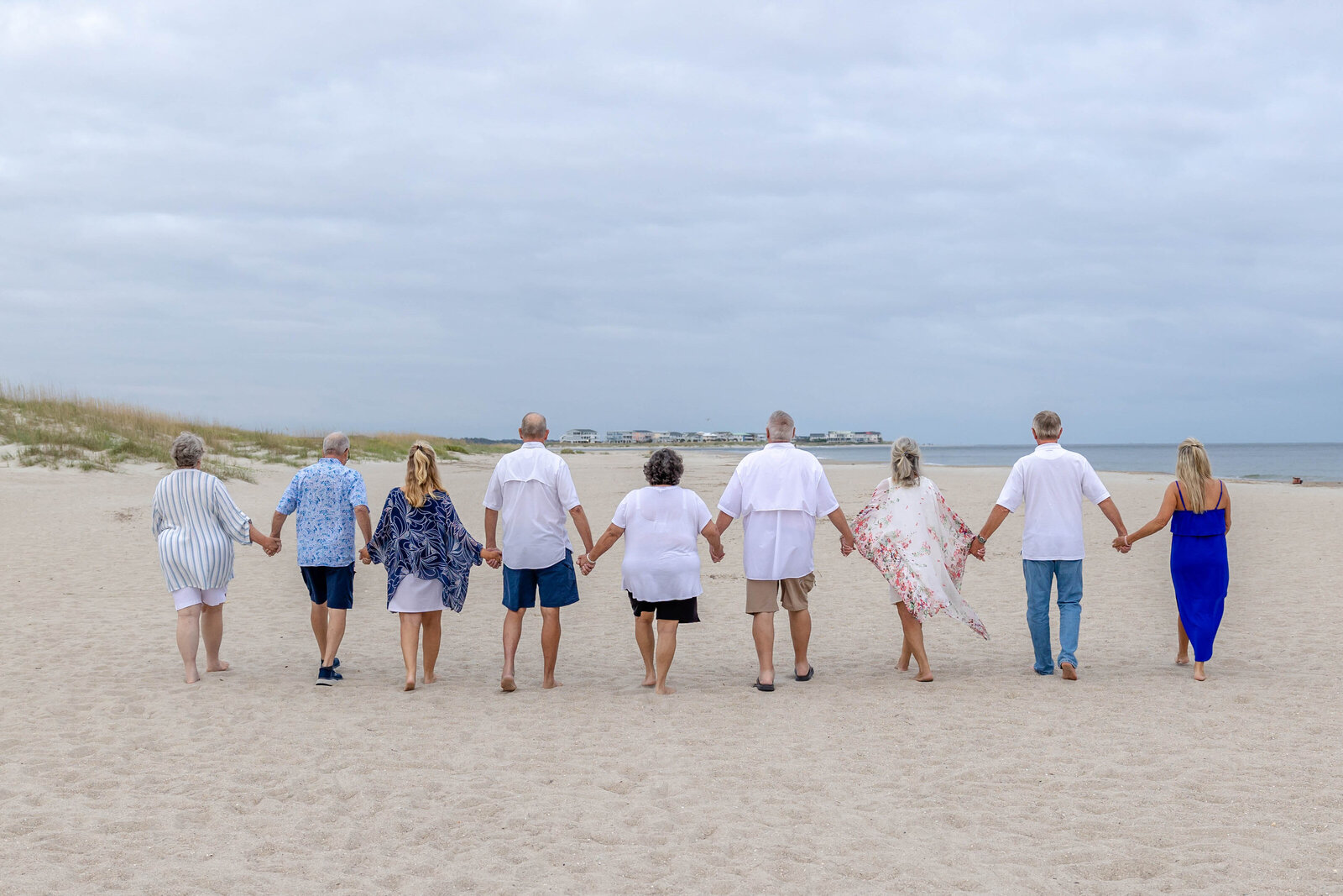 A group walks hand in hand along the sandy beach, dressed in casual summer attire. The overcast sky adds a peaceful touch, ideal for professional family photography in NC, showcasing unity and connection by the sea.