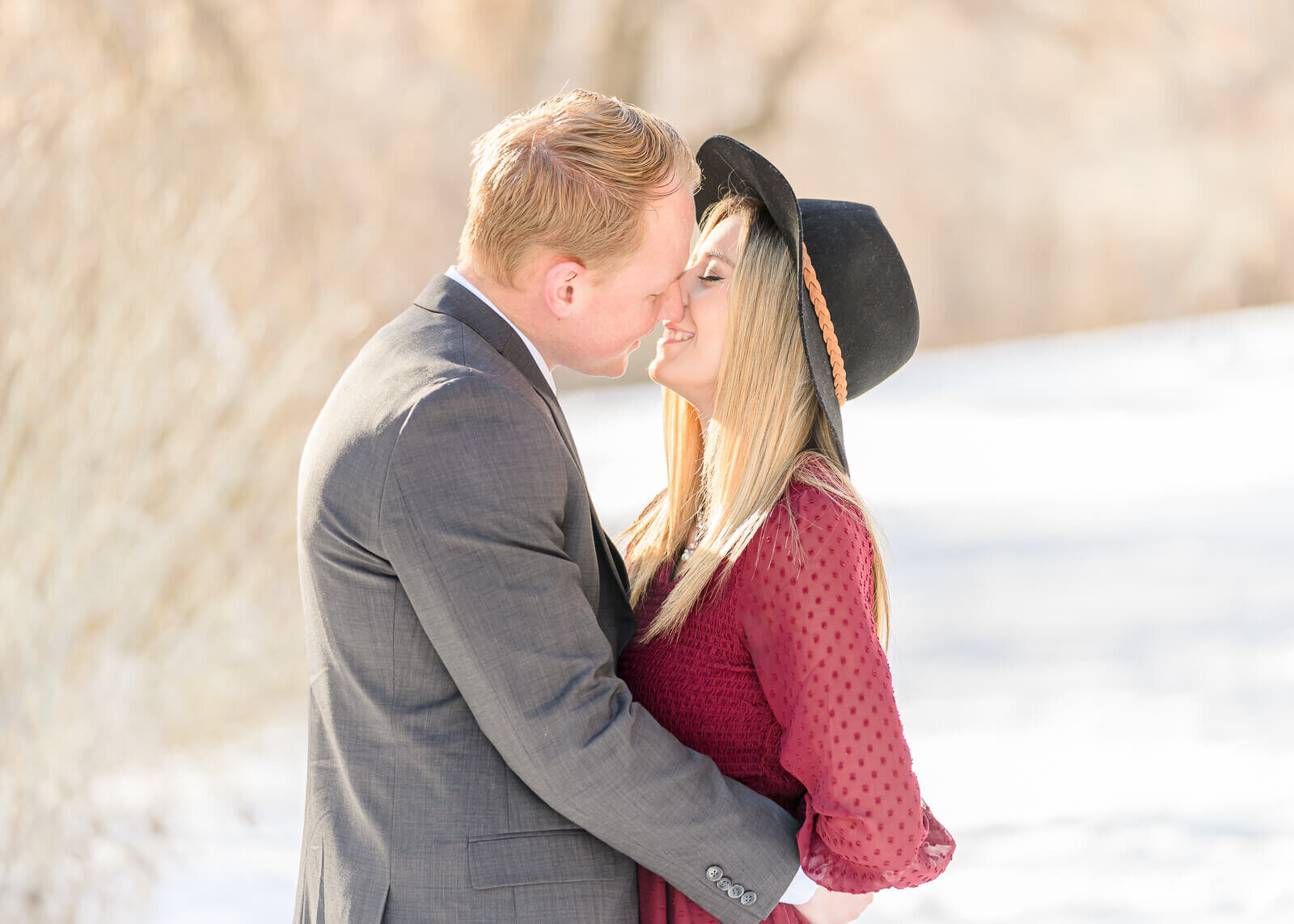 Utah engagement photography of a man in a gray suit leaning in to kiss his fiancee wearing a burgundy long sleeve dress while standing in the snow at Canyon Glen Park, Provo