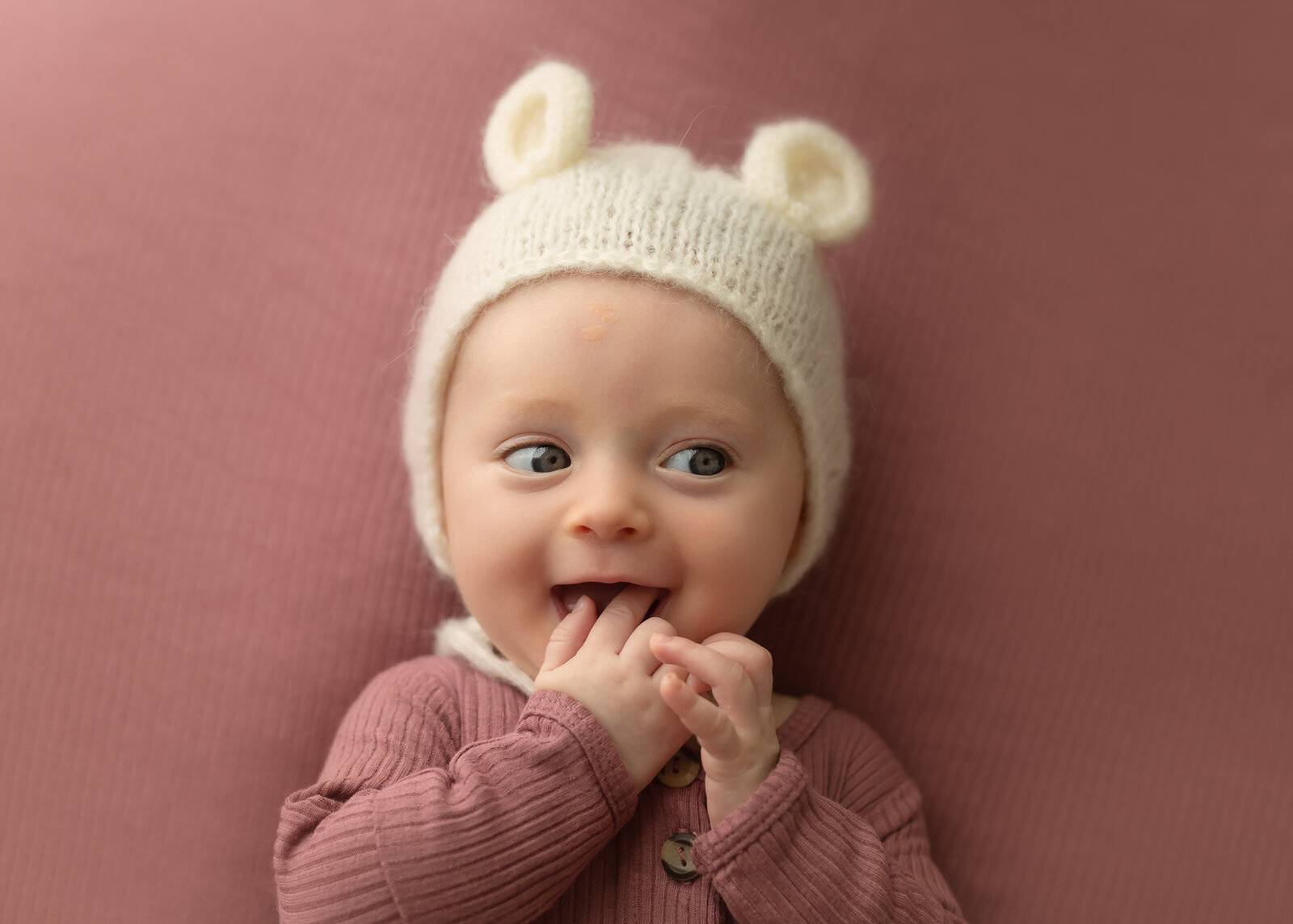 happy baby on mauve background with a teddy bear hat and fingers in mouth