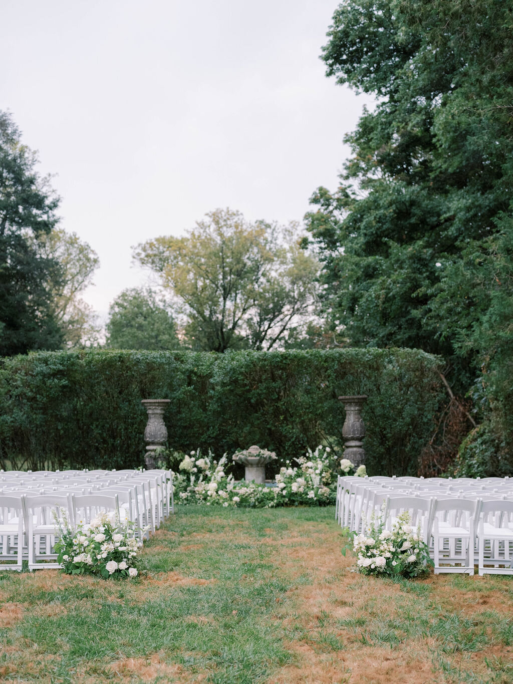 Ceremony in the outside garden at the Evergreen Museum with floral welcome arrangements at the end of the aisle and a lush white floral ground arch framing the water fountain.