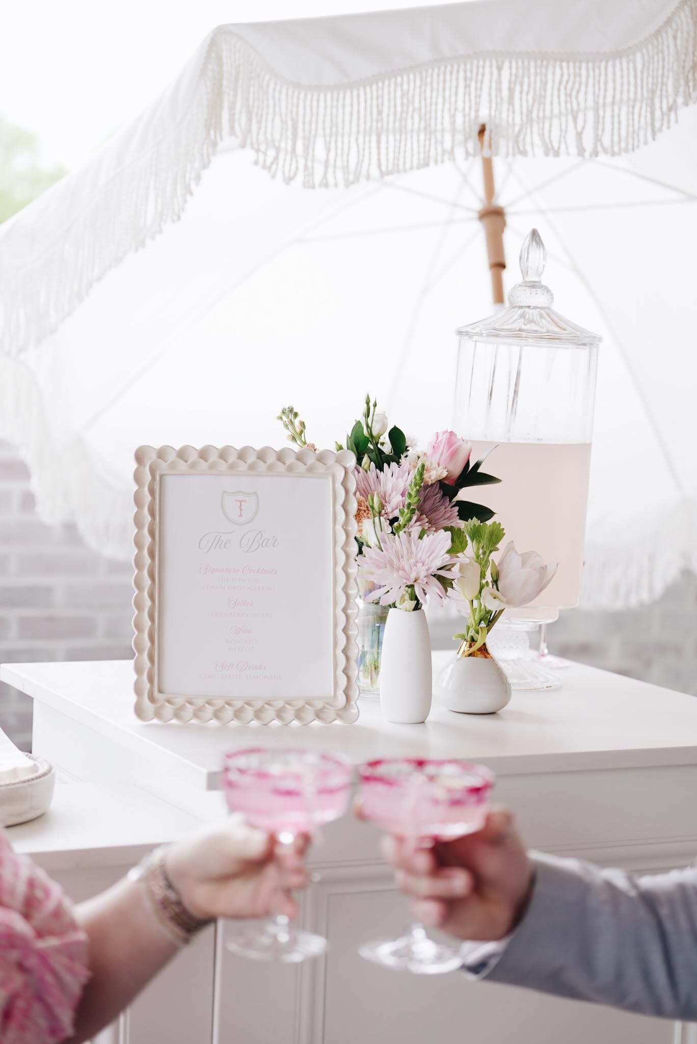 white drink station with pink floral arrangements set under a fringed parasol, with two pink glasses raised in a toast.