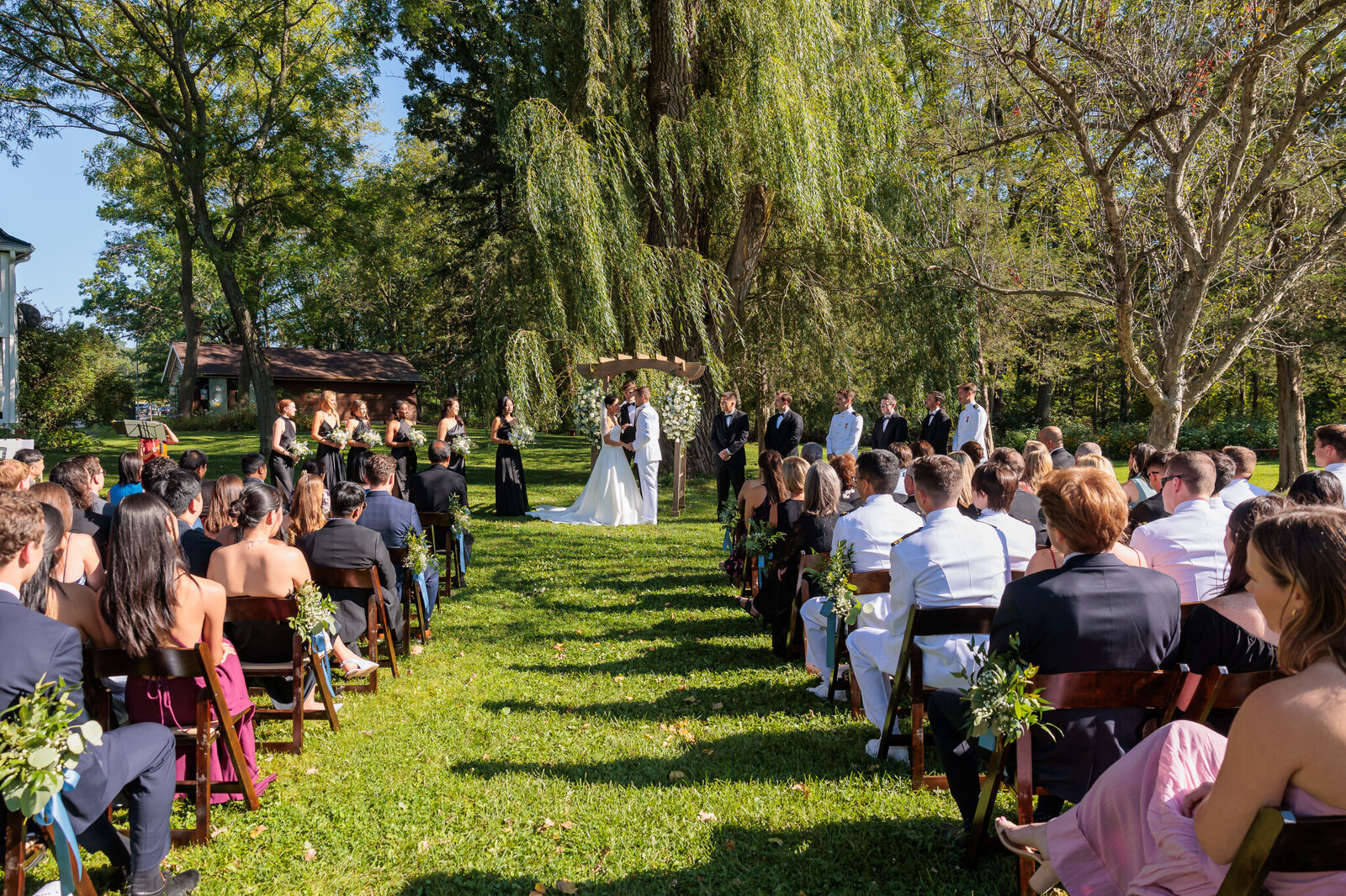 Outdoor wedding ceremony at Barnwood Events in Madison, Wisconsin, with guests seated on the lawn facing the couple under a willow tree.