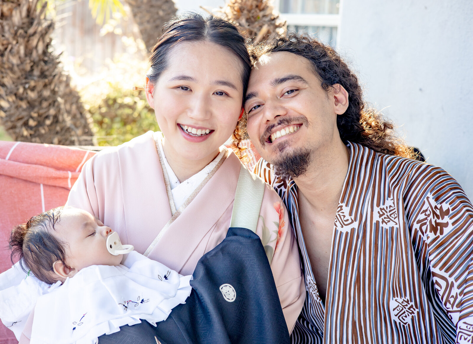 Close-up of smiling parents during a hatsumairi baby blessing in Los Angeles, mother in pink kimono holding baby in black baby kimono and father in brown striped yukata, perfect for kimono rental in Los Angeles | ロサンゼルスで初参りベビー祝をする家族写真のクローズアップ、ピンク着物のお母さんが黒祝い着の赤ちゃんを抱き、茶色ストライプ浴衣のお父さんが微笑む姿。ロサンゼルスの着物レンタルにおすすめ