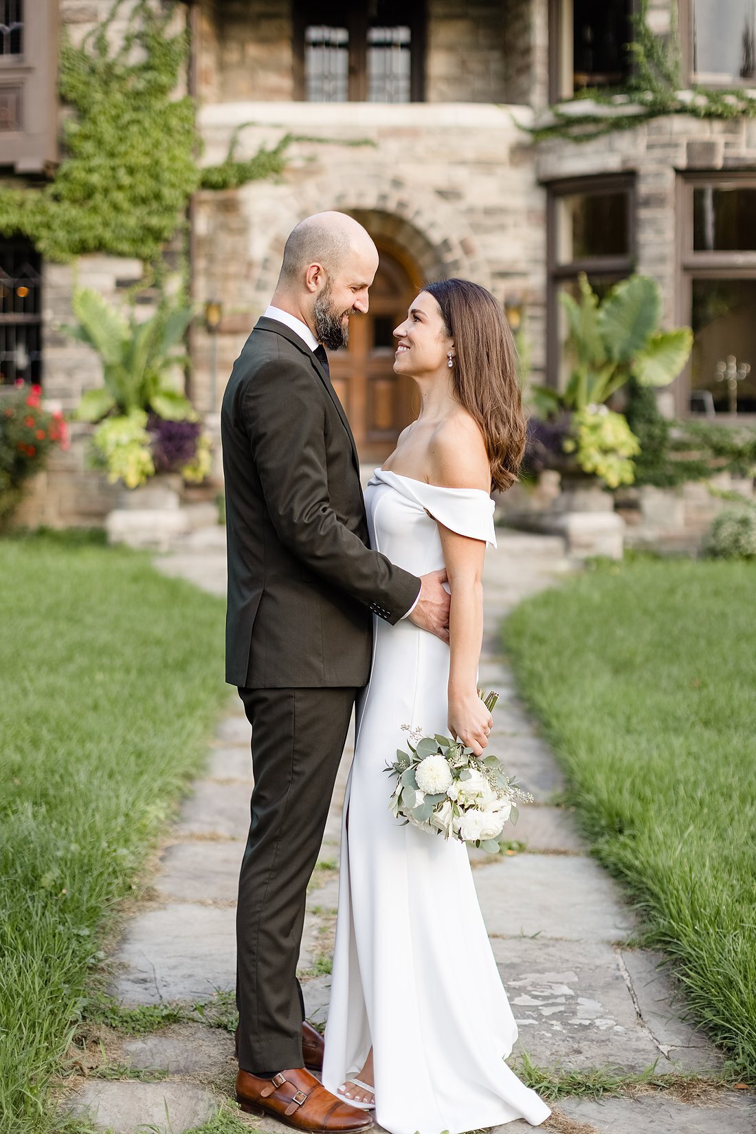 Stylish-groom-embraces-his-stunning-bride-as-they-look-into-each-others-eyes-in-front-of-their-wedding-mansion-in-ontario