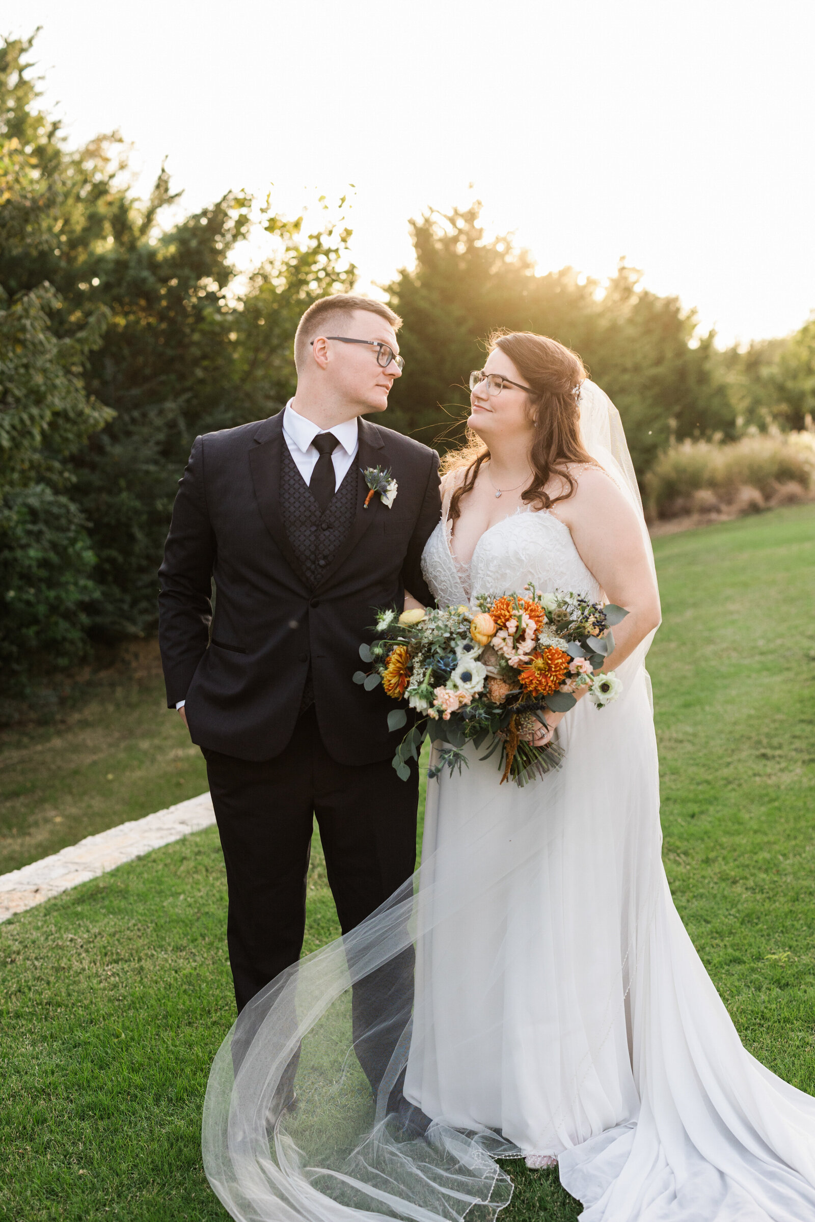 A romantic portrait of a bride and groom outside the Laurel Grapevine, Texas. The bride is on the right and is wearing a white dress with a long flowing veil and is holding a large bouquet. The groom is on the left and is wearing a black suit with a boutonniere.