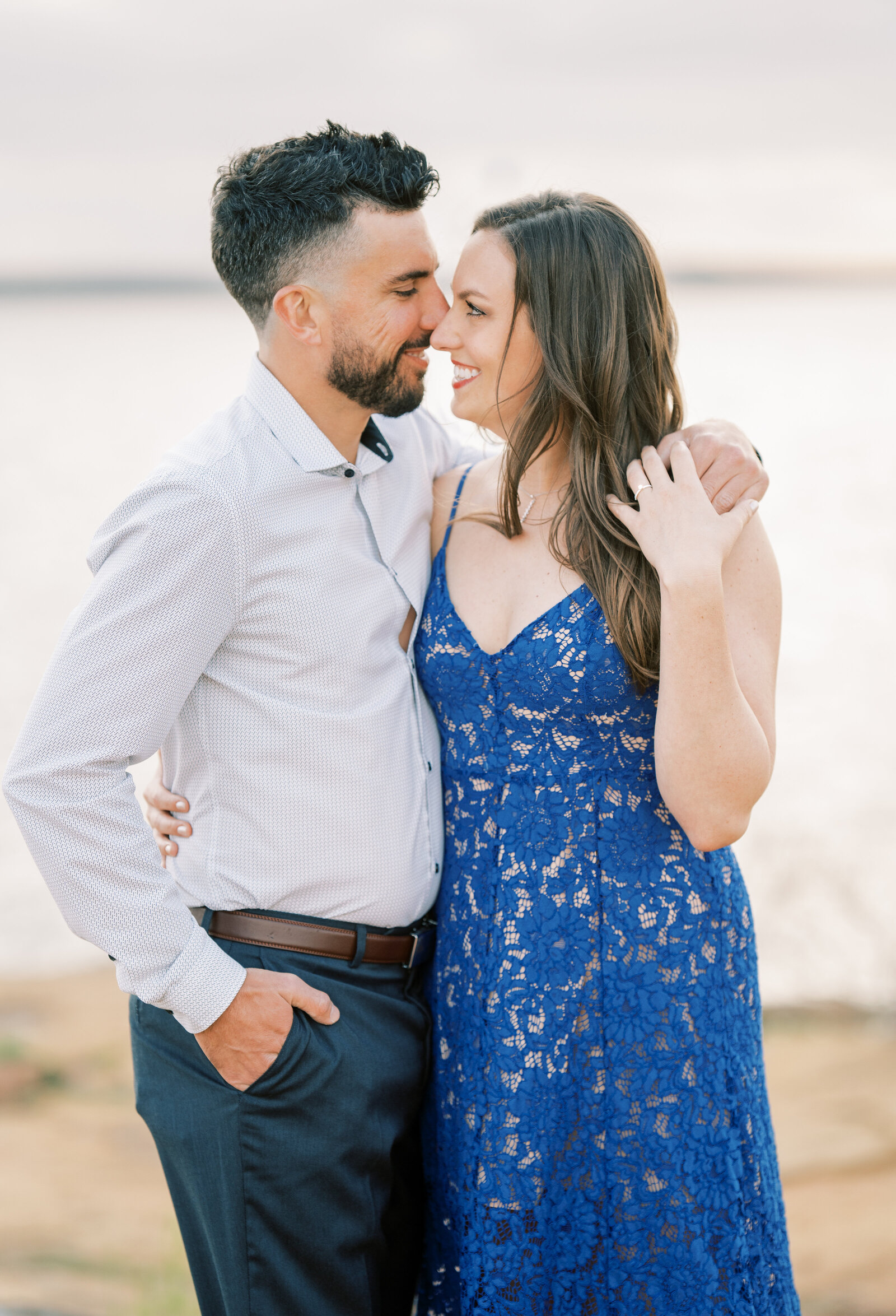 Portrait of a man in a white shirt and dress pants hugging a woman in a blue lace dress while standing on sand at the beach