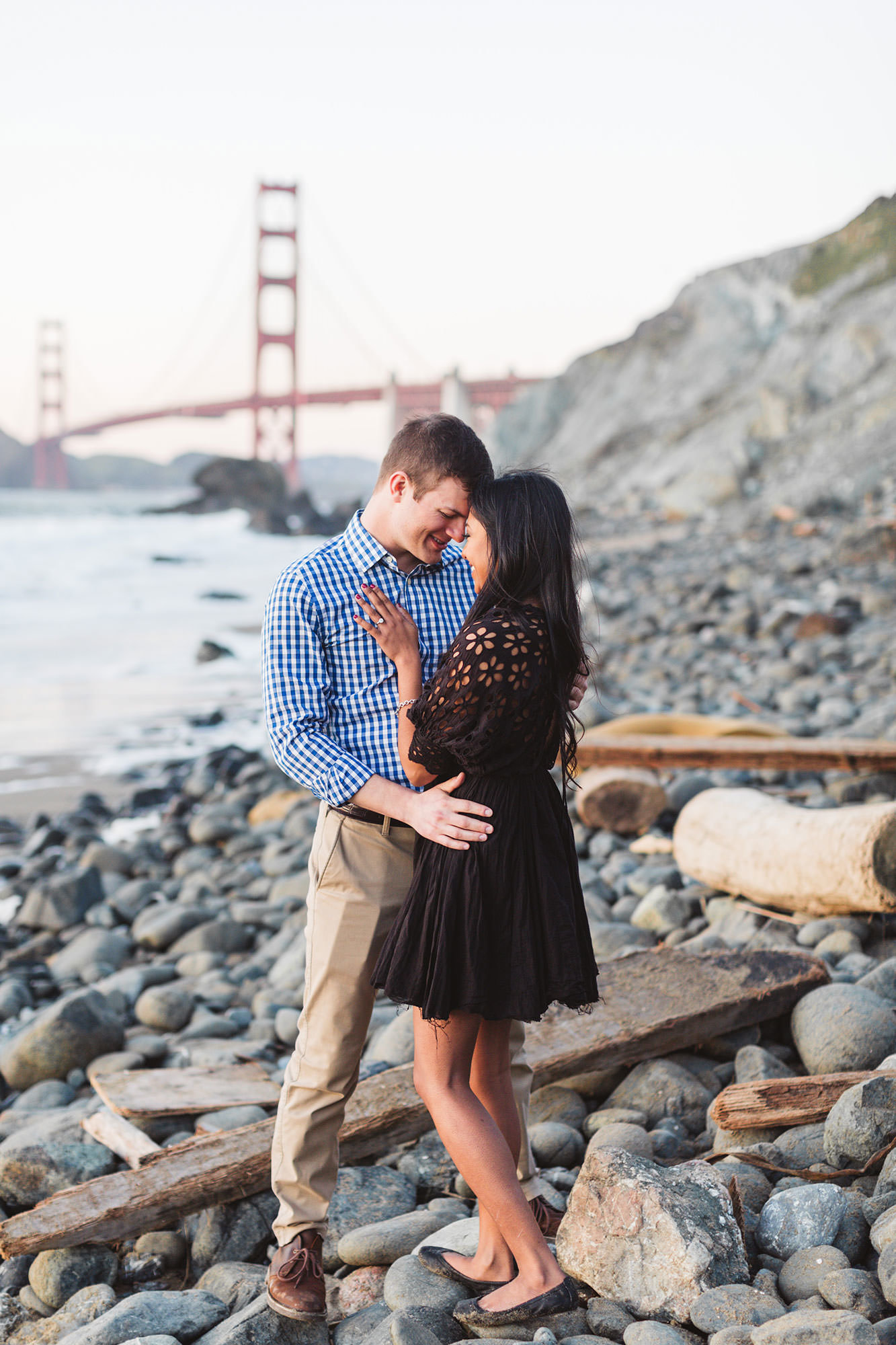 engaged couple putting foreheads together on marshall's beach with golden gate bridge beindd