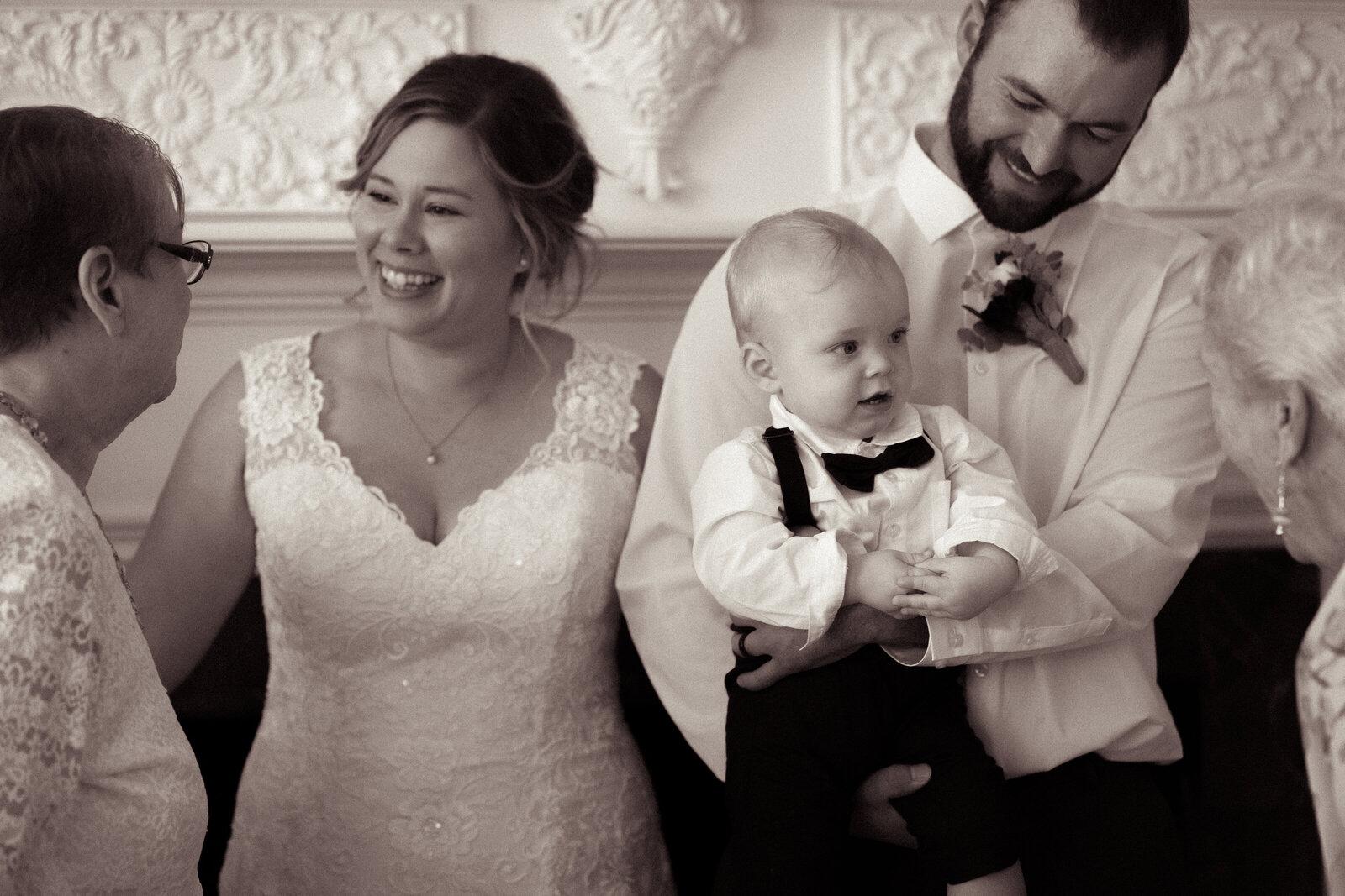 cinematic, relaxed photo of groom holding son, who's looking at grandma, while bride talks to a guest.