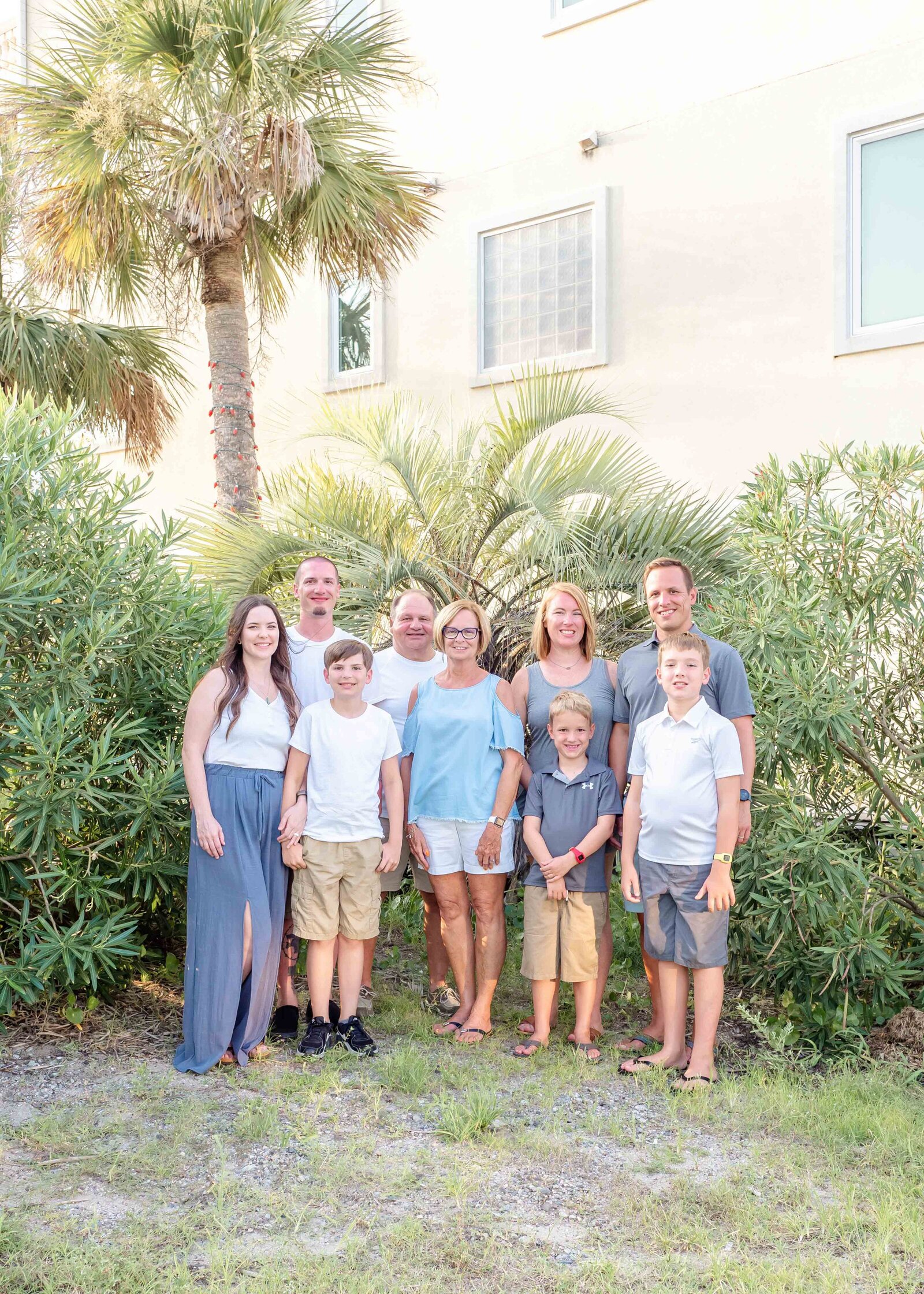 A multi-generational family stands together by palm trees, dressed in casual summer clothes. The bright greenery and relaxed expressions highlight their close-knit bond. Perfect for professional family photography in NC, showcasing family unity and happiness.