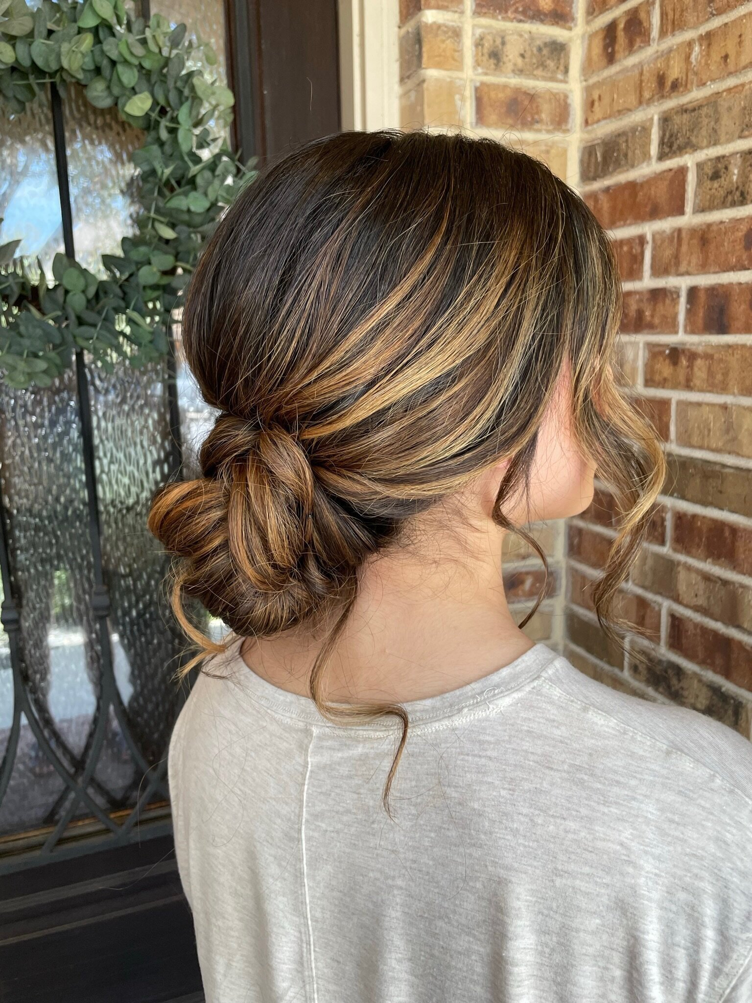 A woman with highlighted hair styled in a braided low bun stands by a brick wall and glass door with a wreath, embodying the essence of bridal beauty experience in Texas.