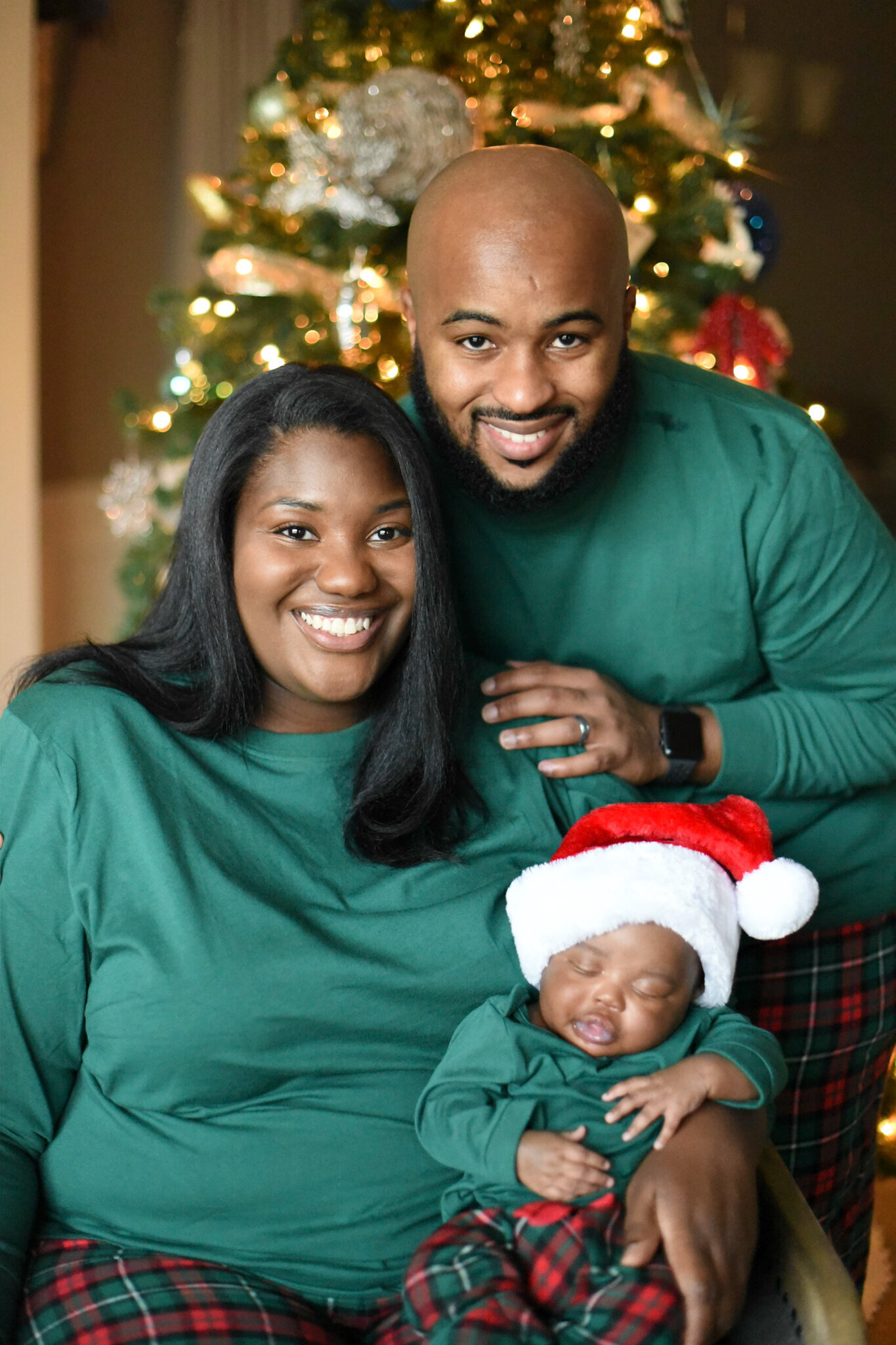a mother and father smiling in front a a christmas tree with their newborn baby who is celebrating his first christmas. they are all wearing matching pajamas photographed by Millz Photography in Greenville, SC