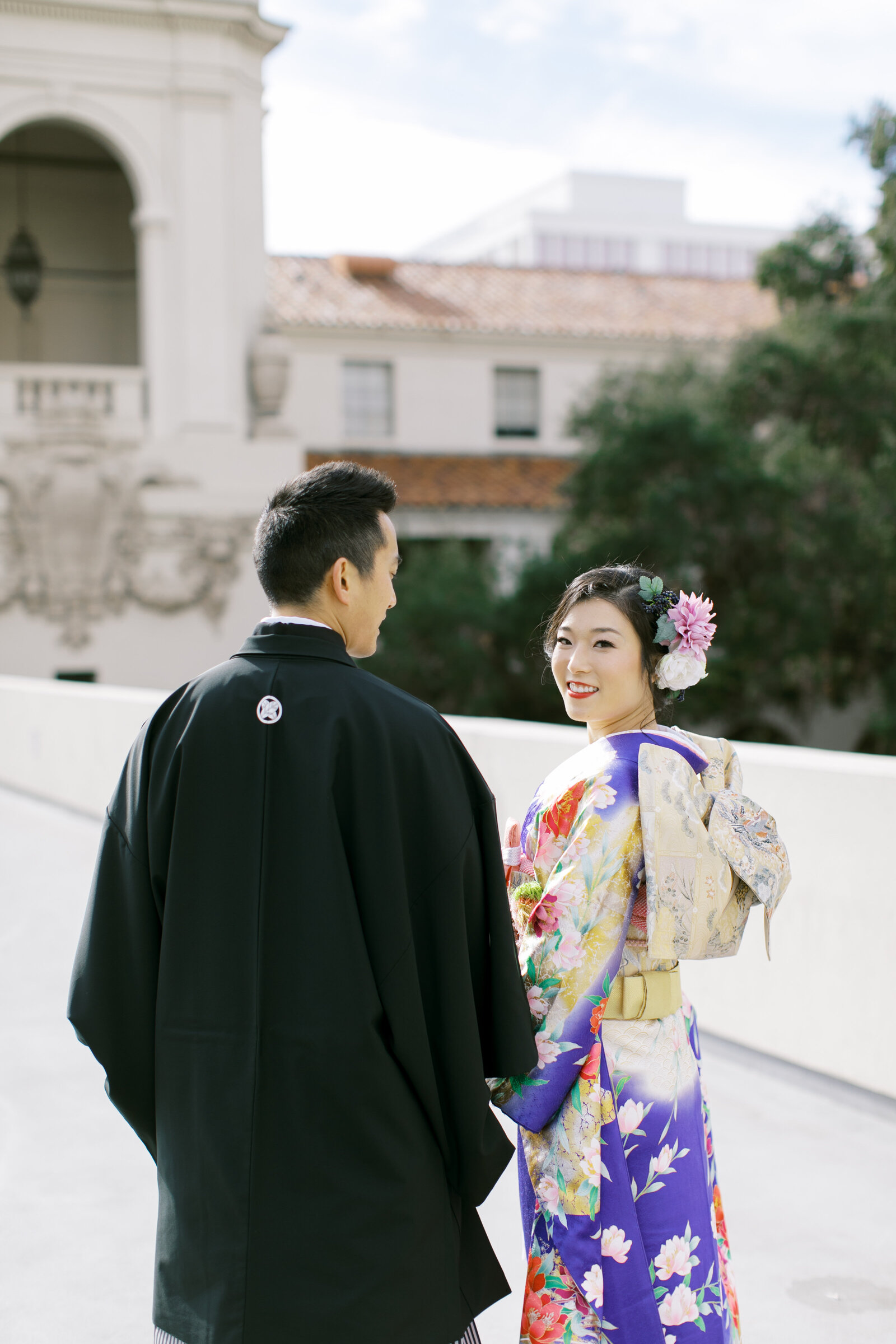 Bride in purple floral wedding kimono rental glancing back at groom in black montsuki as they walk near Pasadena City Hall in Los Angeles | パサデナ市庁舎周辺を歩く着物レンタルウェディング 紫色振袖の花嫁と黒紋付の新郎