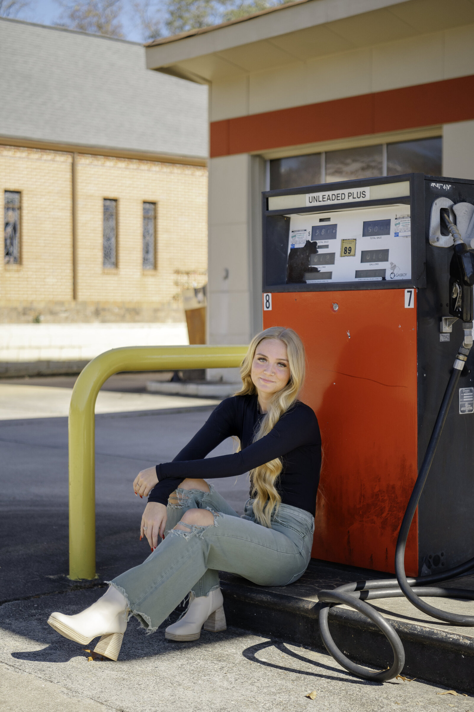 Blonde senior sits beside old red gas pump and looks into the camera during her senior session