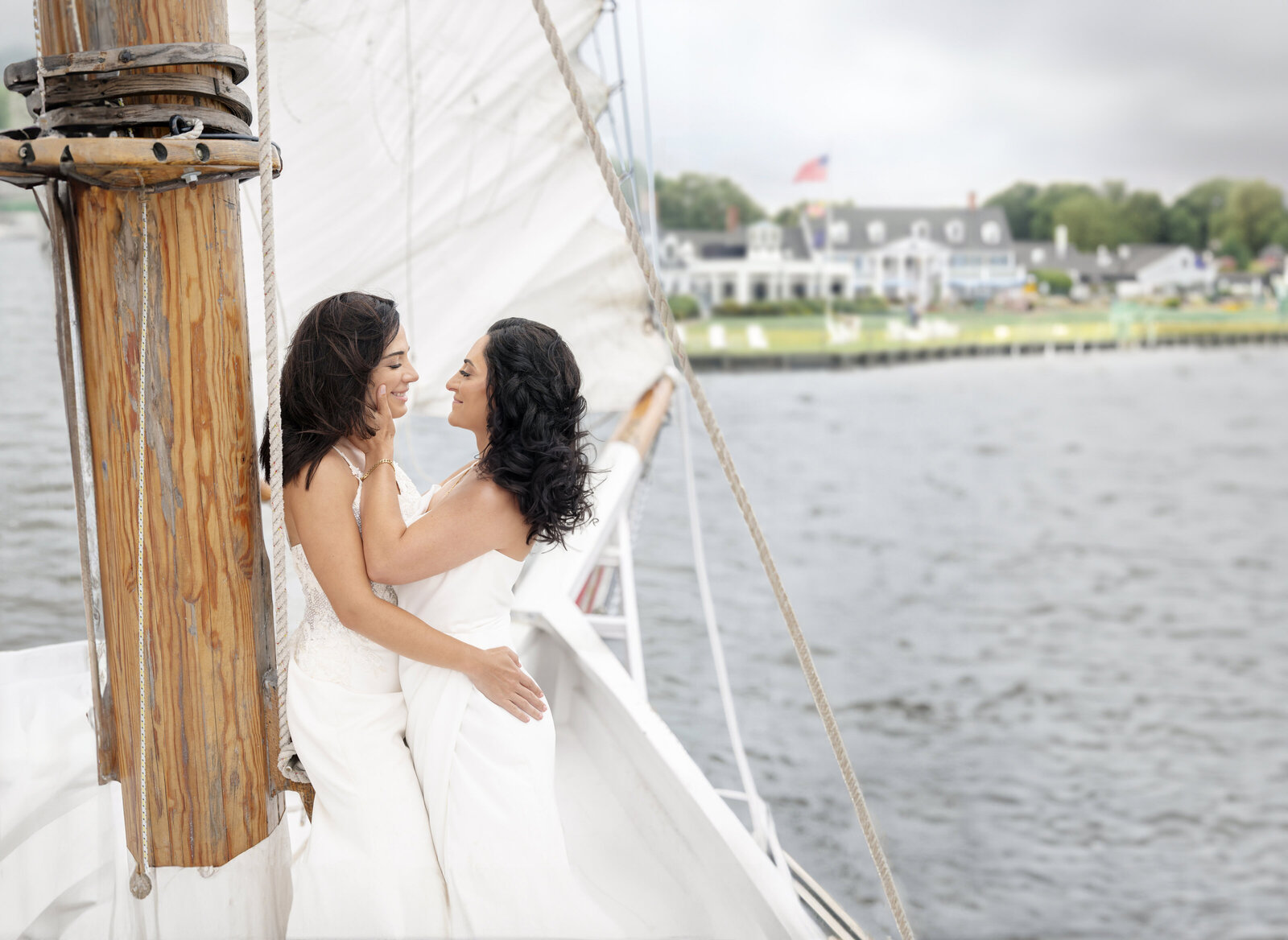 Two women in white dresses embrace on a sailboat, smiling at each other. The boat sails on a body of water with a view of land, a house, and a flag in the background under a cloudy sky.