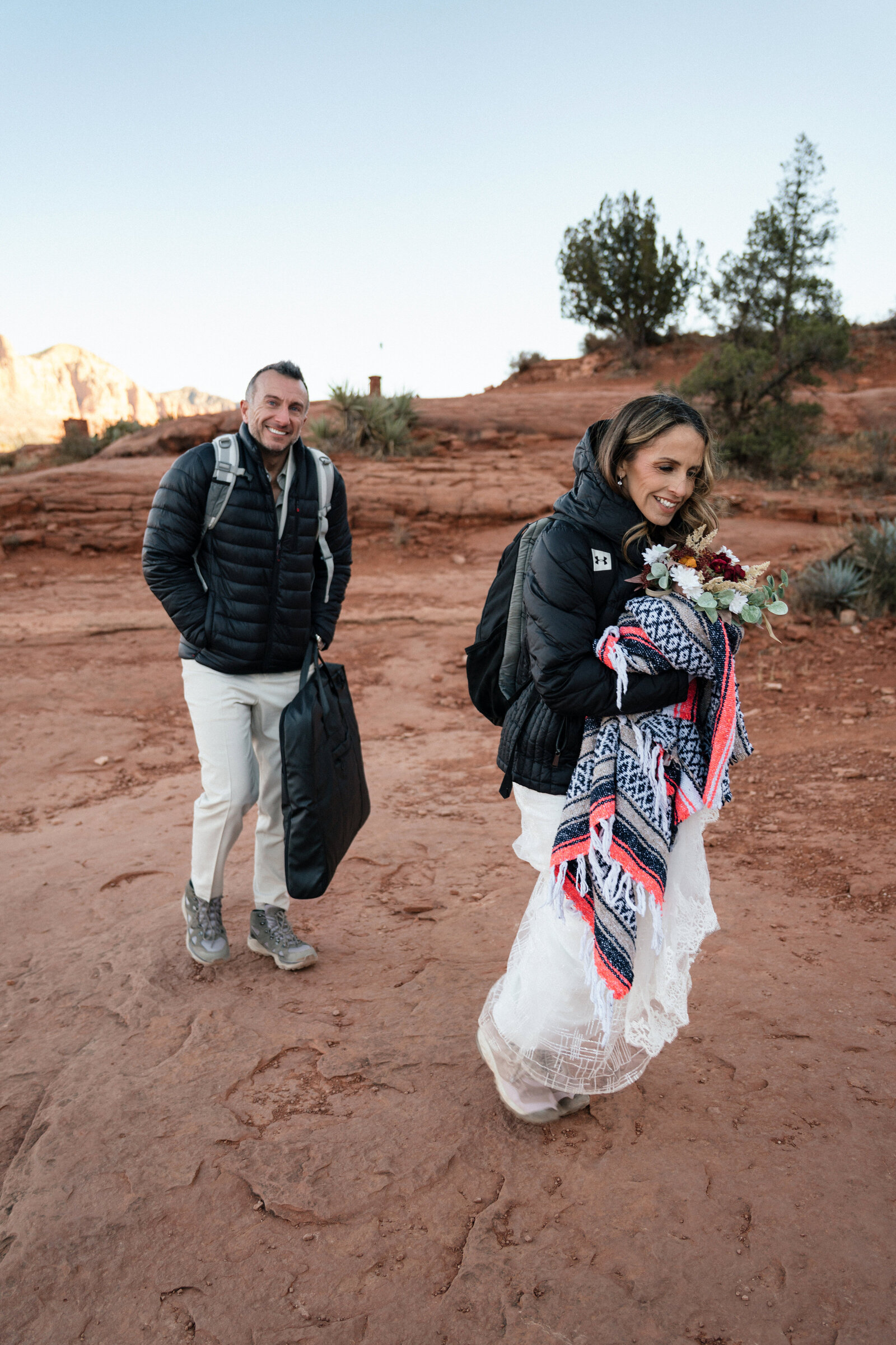 Bride and groom hiking to their elopement at Cathedral Rock in Sedona, Arizona, dressed warmly with backpacks, a bouquet, and a blanket, showcasing a real adventure wedding experience.