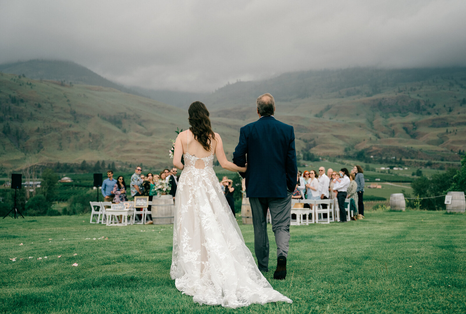 a bride walking towards her wedding ceremony holding her dad's arm in front of green mountains with stormy moody skies