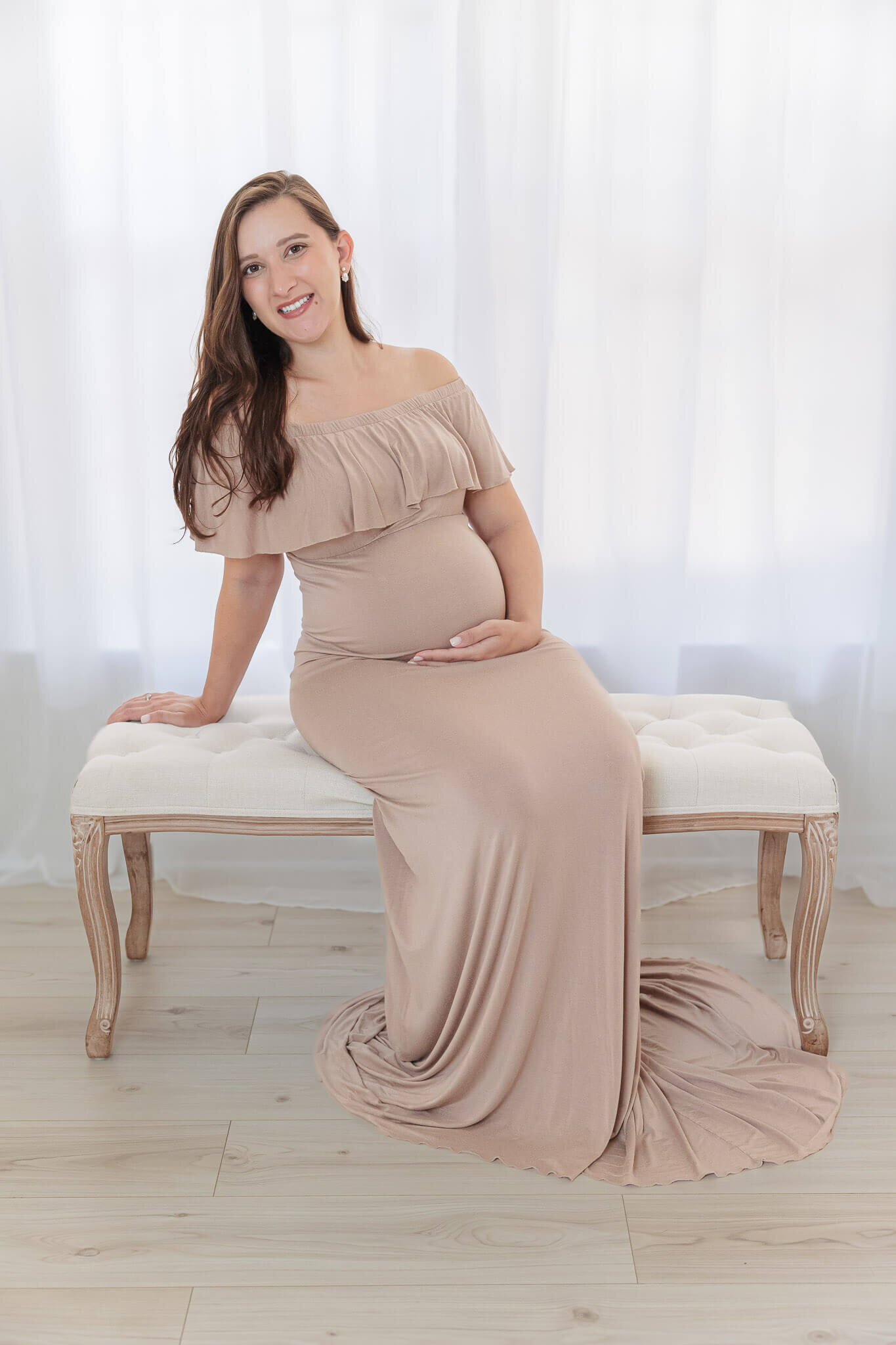 A pregnant mom in a tan dress sitting on a bench in front of a window.