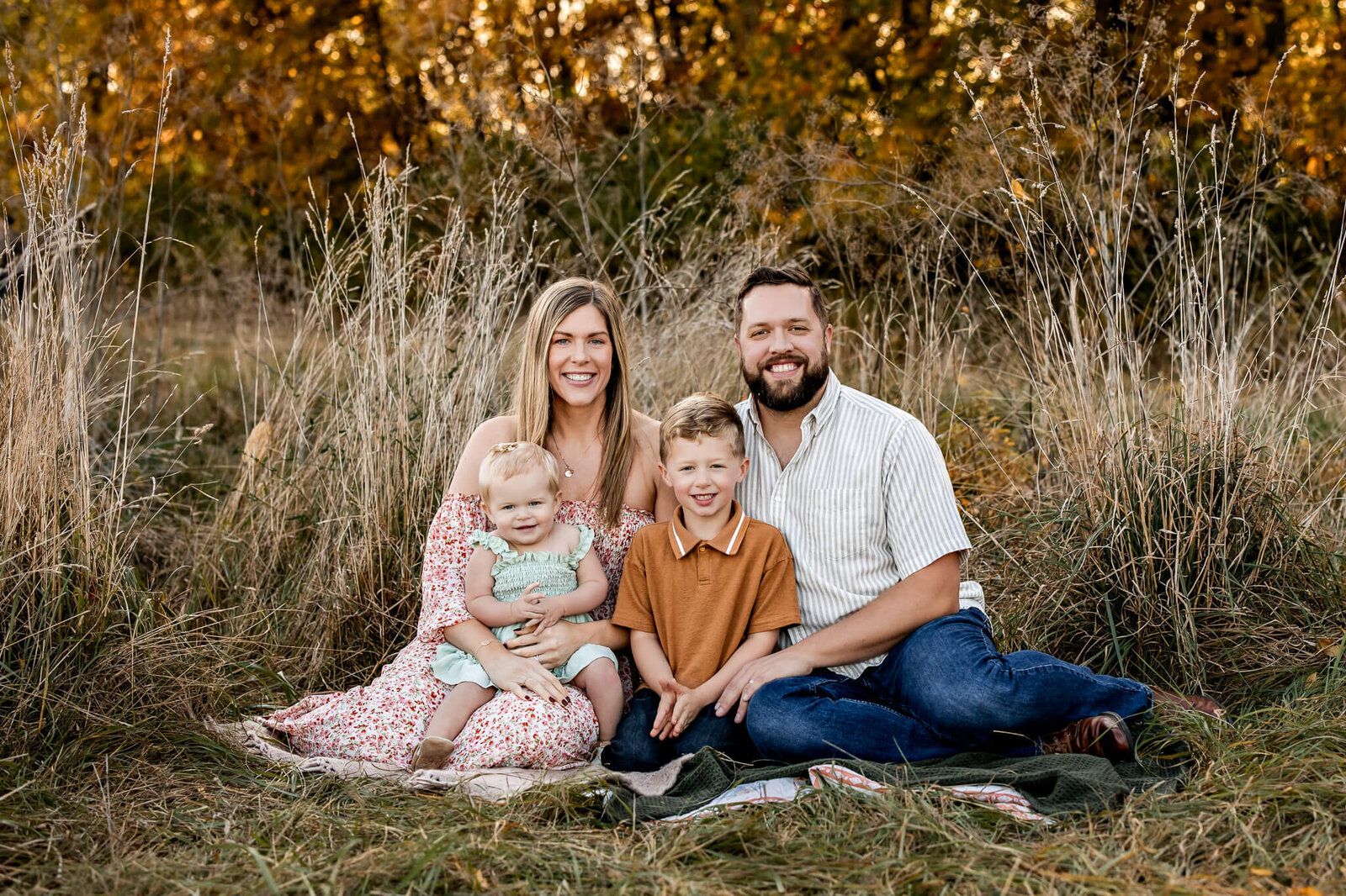 A Bloomington family photographer captures a vibrant outdoor portrait of a family of four, seated on a picnic blanket in a grassy field adorned with autumn foliage. The mother holds a toddler in a dress, while the father sits beside their young boy in a polo shirt. Smiles abound as they face the camera.