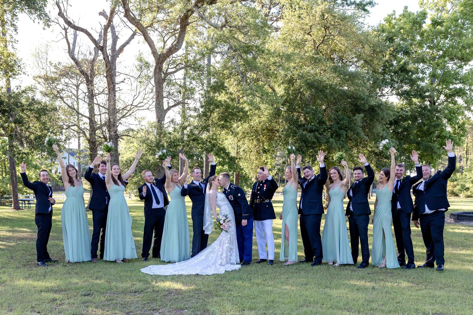 A wedding party dressed in sage green and navy attire celebrates with the bride and groom on a sunny lawn at a Wrightsville Beach, NC wedding, blending joy and coastal elegance.