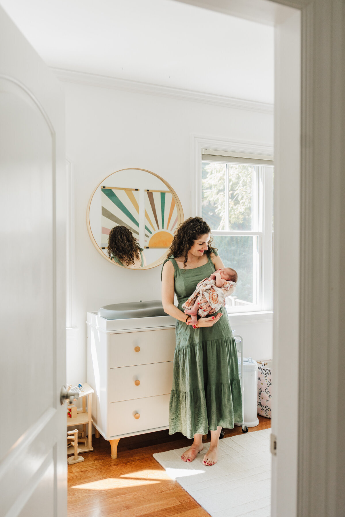 mom in green dress leans against changing table in nursery while gazing at newborn daughter