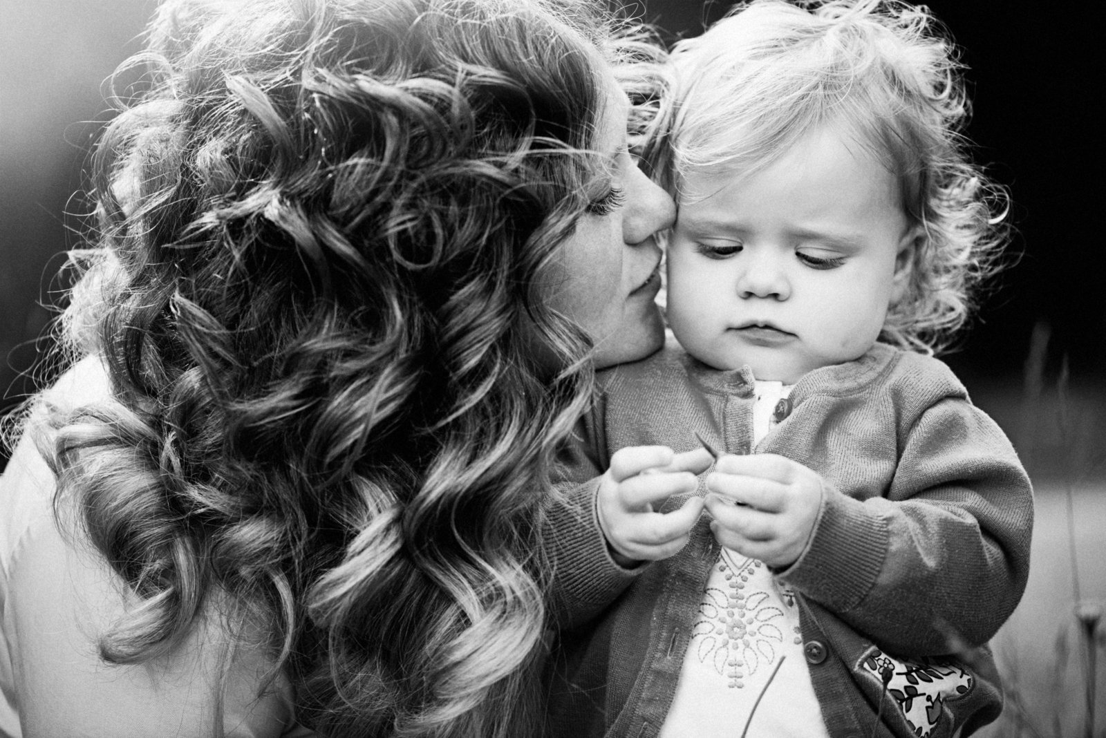 A black and white picture of a toddler being kissed on the cheek by her curly haired mother