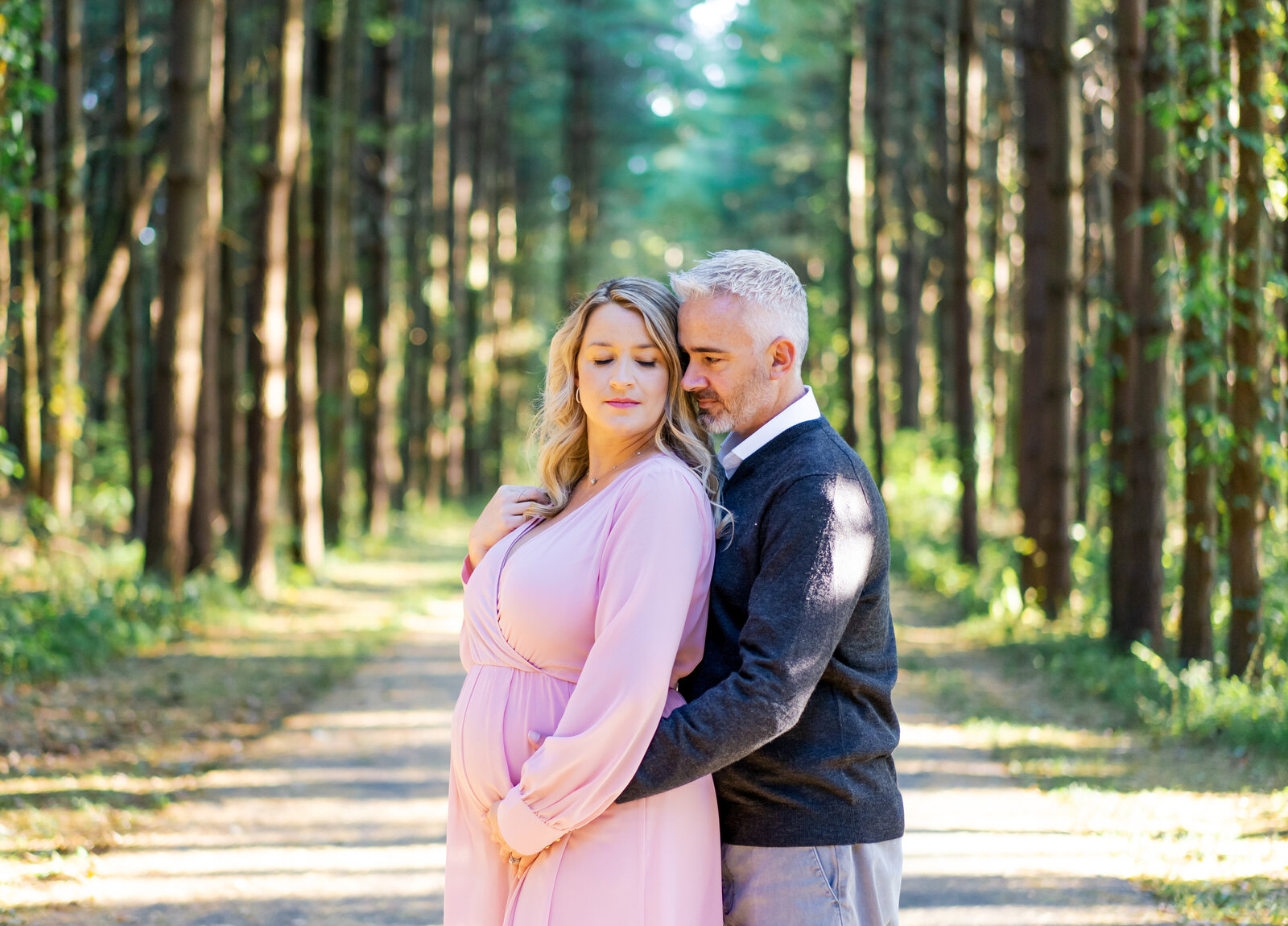 Husband holding his pregnant wife while she holds her belly and looks off her shoulder at Walnut Woods Tall Pines in Groveport, Ohio. Maternity photo.