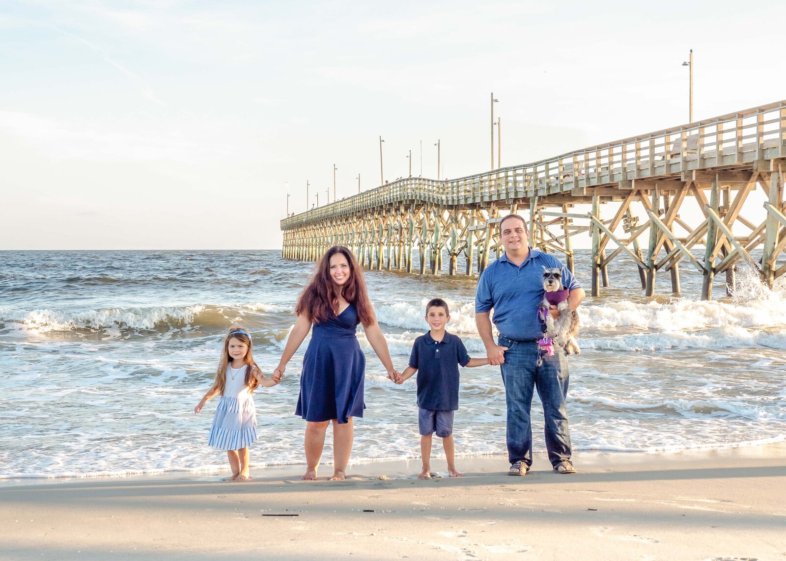 A family of four holds hands on the beach near a pier, with their dog in a festive outfit. Dressed in blue and white, they enjoy a sunny day by the waves. Ideal for professional family photography in NC, highlighting playful seaside moments.