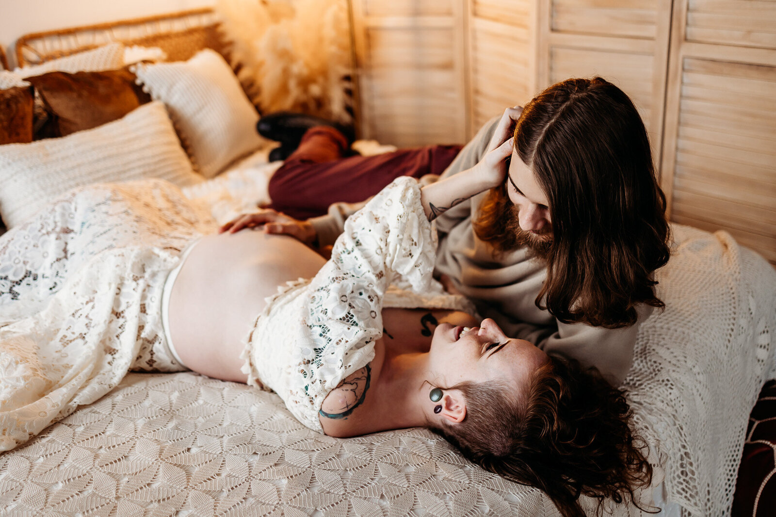 Laying together on a bed surrounded by boho decor, an expecting mother caresses her husband as her holds her pregnant belly