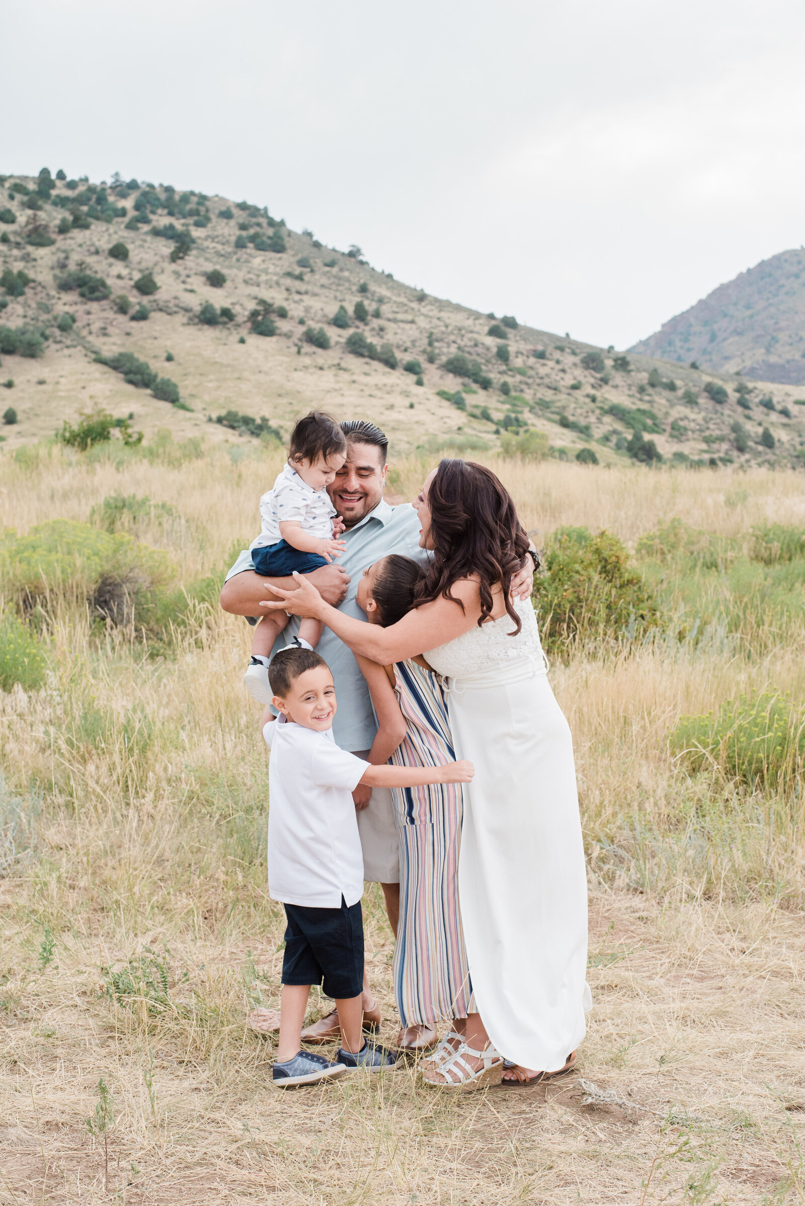 dad holds a toddler boy as the mom and other siblings come in and hug them in a field at the base of the rocky mountains for their family outdoor photos with denver family photographers