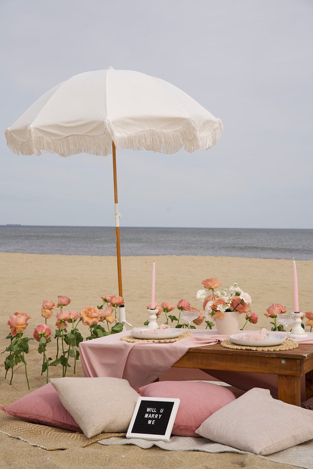 Romantic beach picnic with blush pink florals, elegant table settings, and a white fringe umbrella, styled by Yours Truly, a wedding planner and designer in New Jersey.