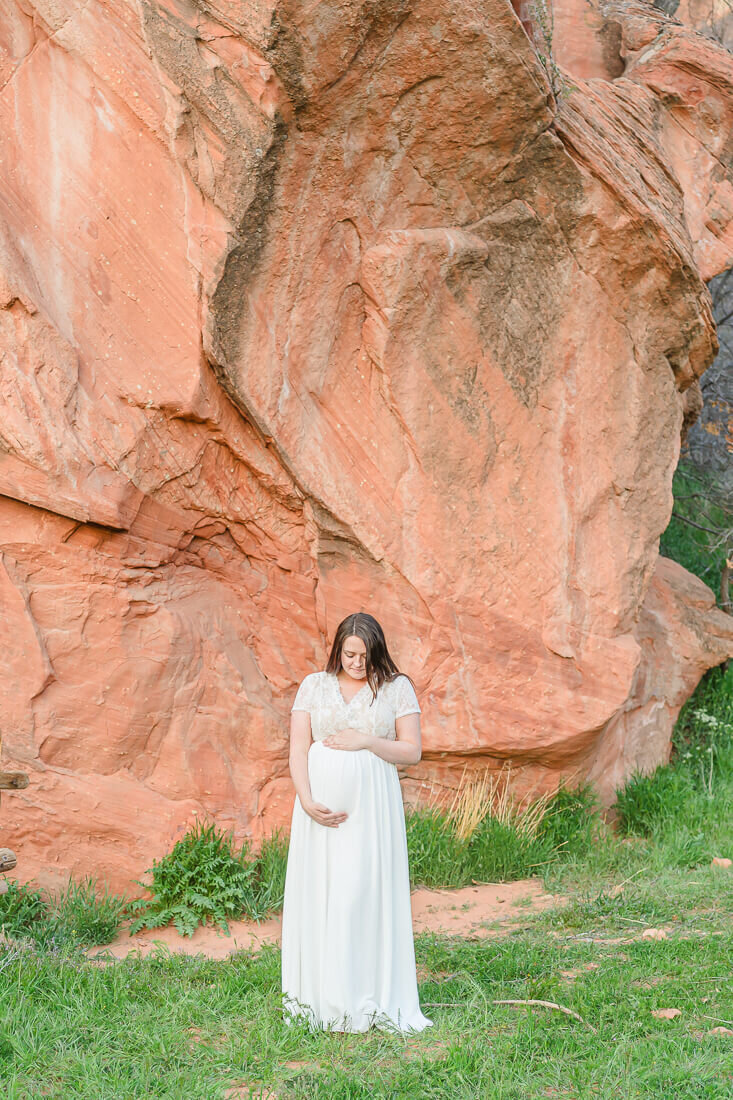 Utah maternity photography of a pregnant woman wearing a long white maternity dress standing in front of the red cliffs at Red Ledges Picnic Area in Spanish Fork
