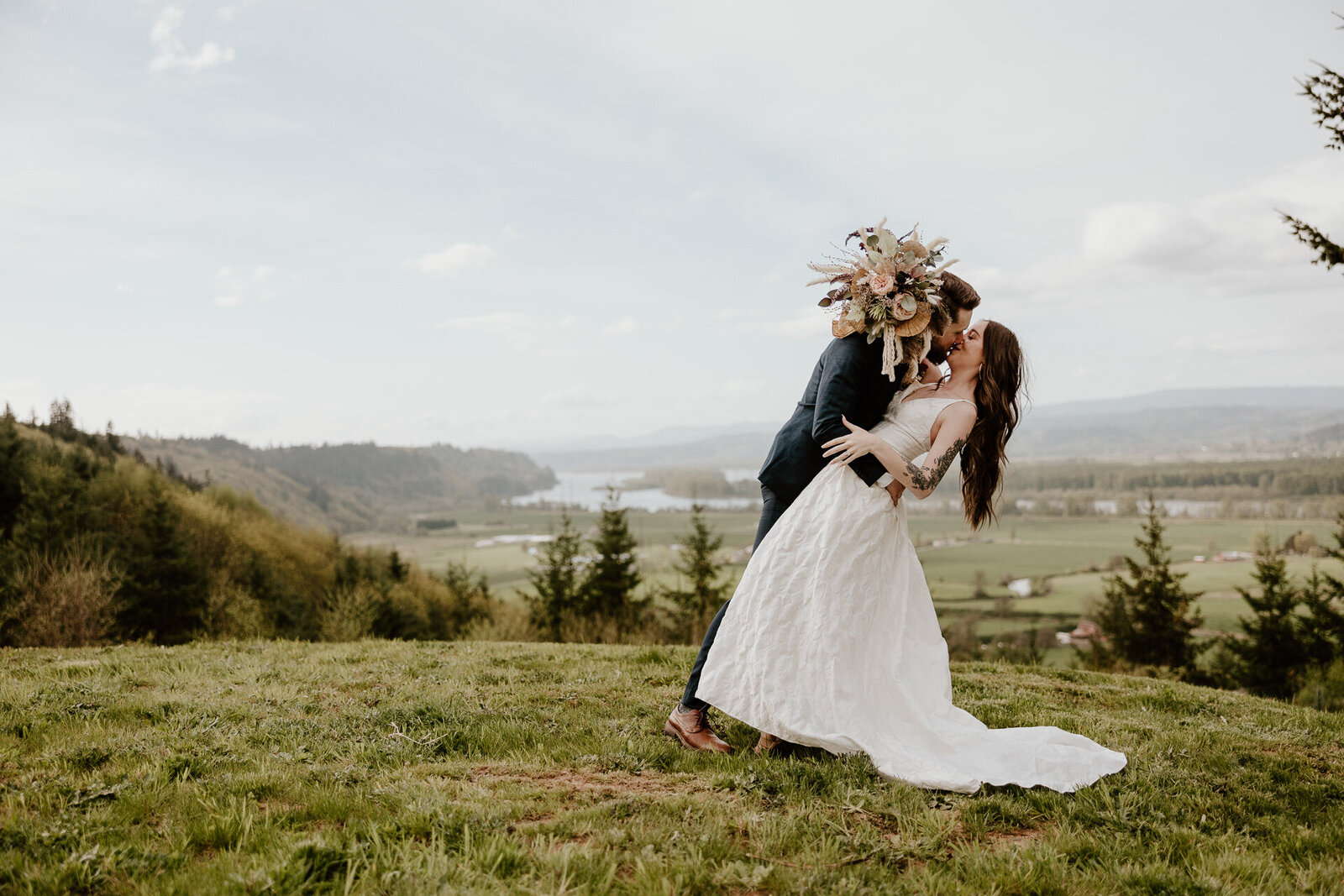 An image captured by a Vancouver WA Wedding Photographer of newlyweds kissing and dipping on top of a hill with a large bouquet