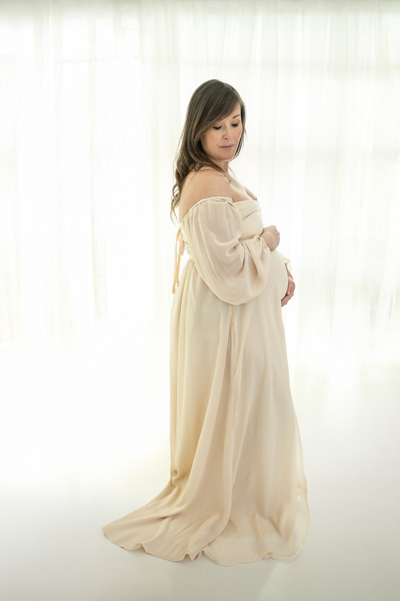 Side profile of expectant mother in off-shoulder dress, softly lit in a minimalist Baltimore photography studio.
